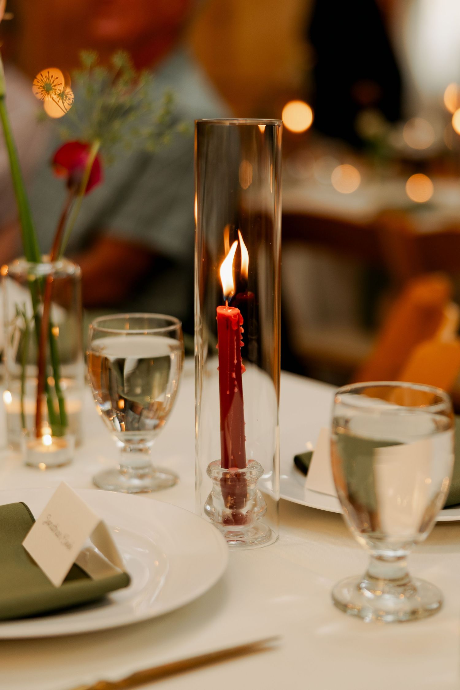 Close-up of a lit red taper candle in a glass cylinder beside water goblets, a place card, and an olive green napkin.