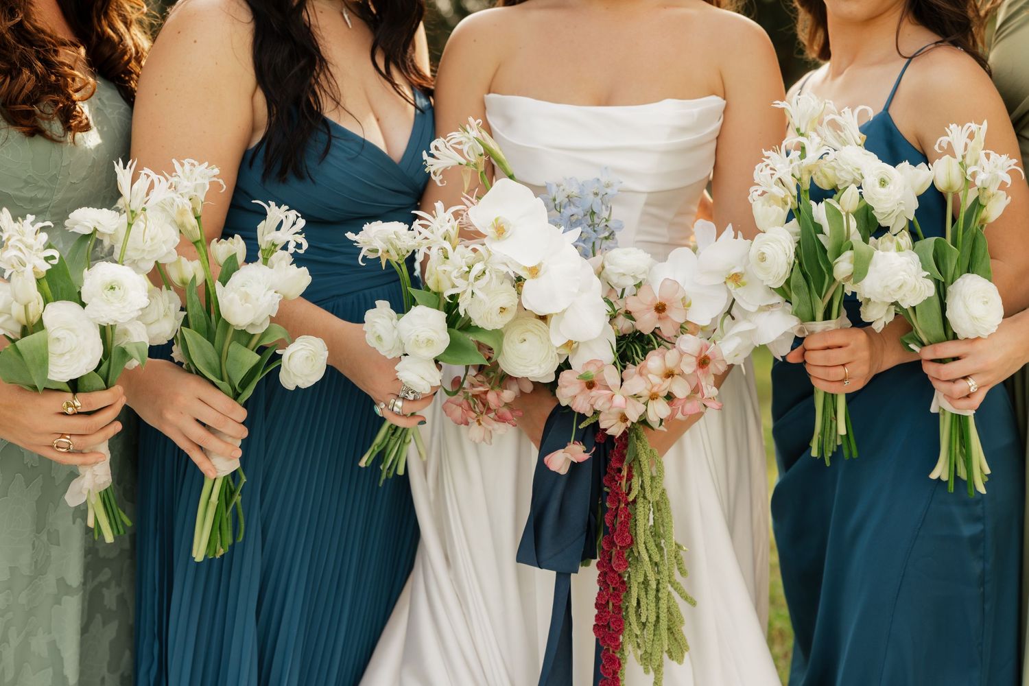 Bride in a white gown and bridesmaids in teal dresses hold white tulip and floral bouquets with greenery at a wedding.