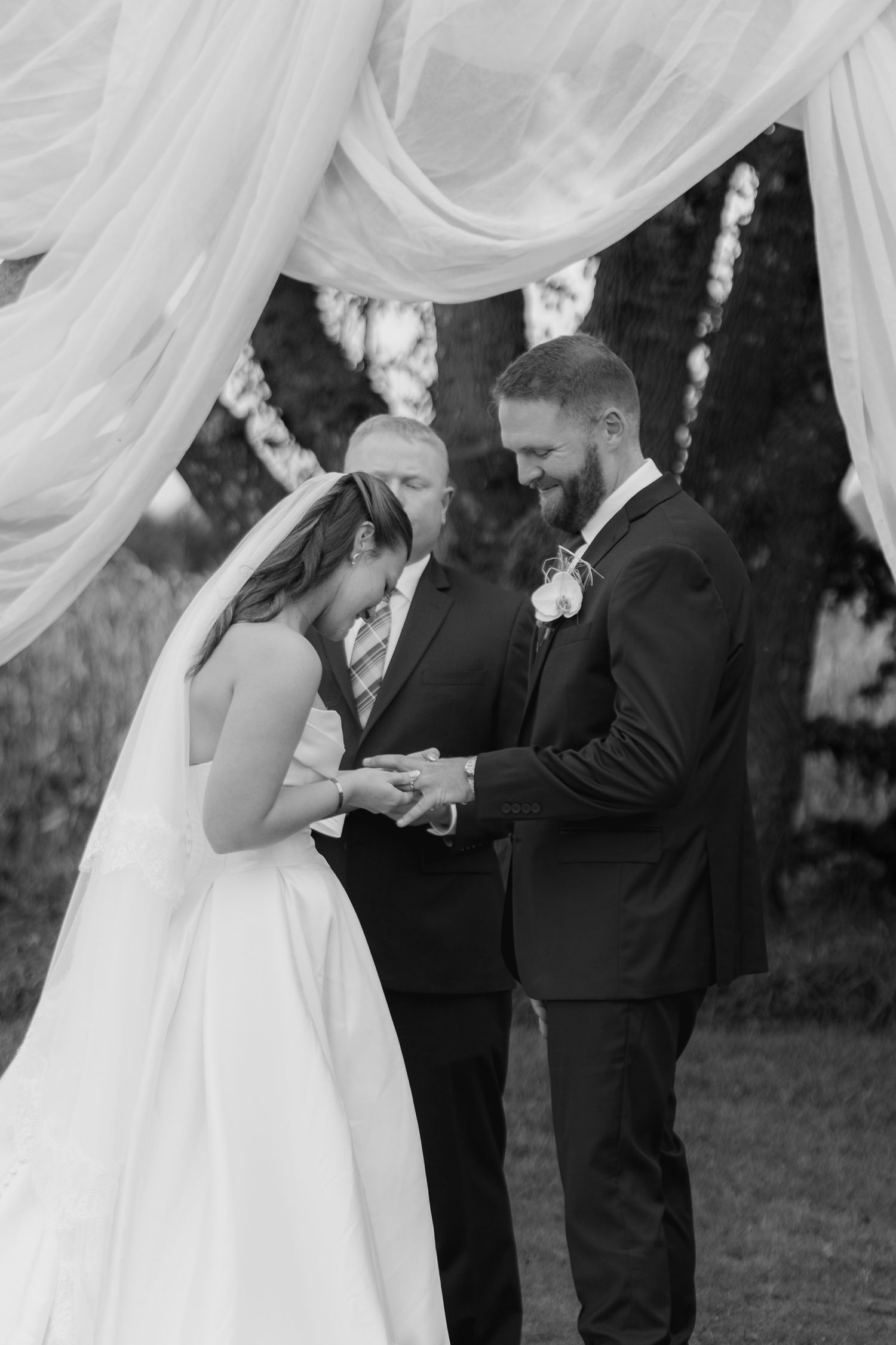 Black and white photo of a bride in a veil and a smiling groom holding hands during their wedding ceremony under draped fabric.