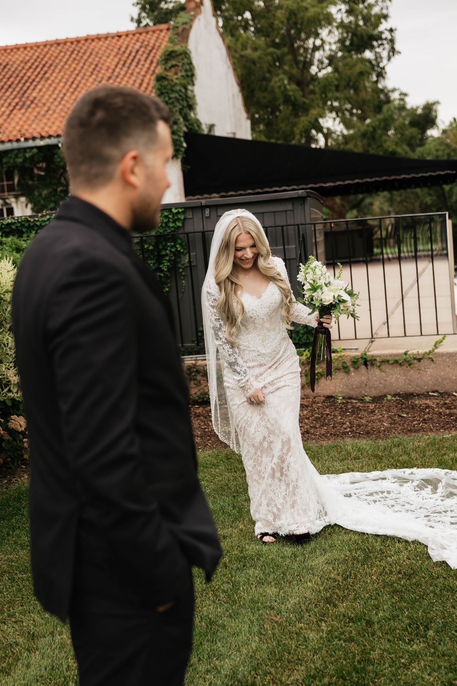Bride in a lace wedding gown and veil walks across a lawn holding a white bouquet toward a groom in a black suit.