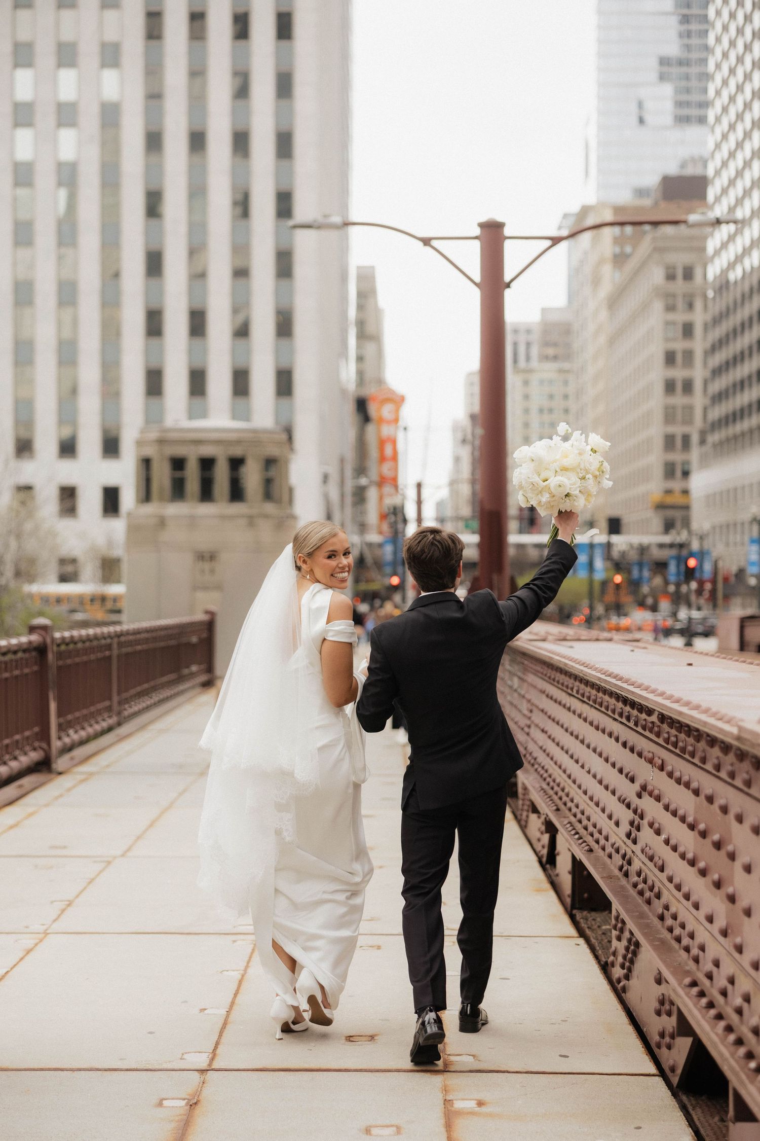 Bride and groom walking on a Chicago bridge, groom raising white bouquet in celebration, city skyline backdrop.