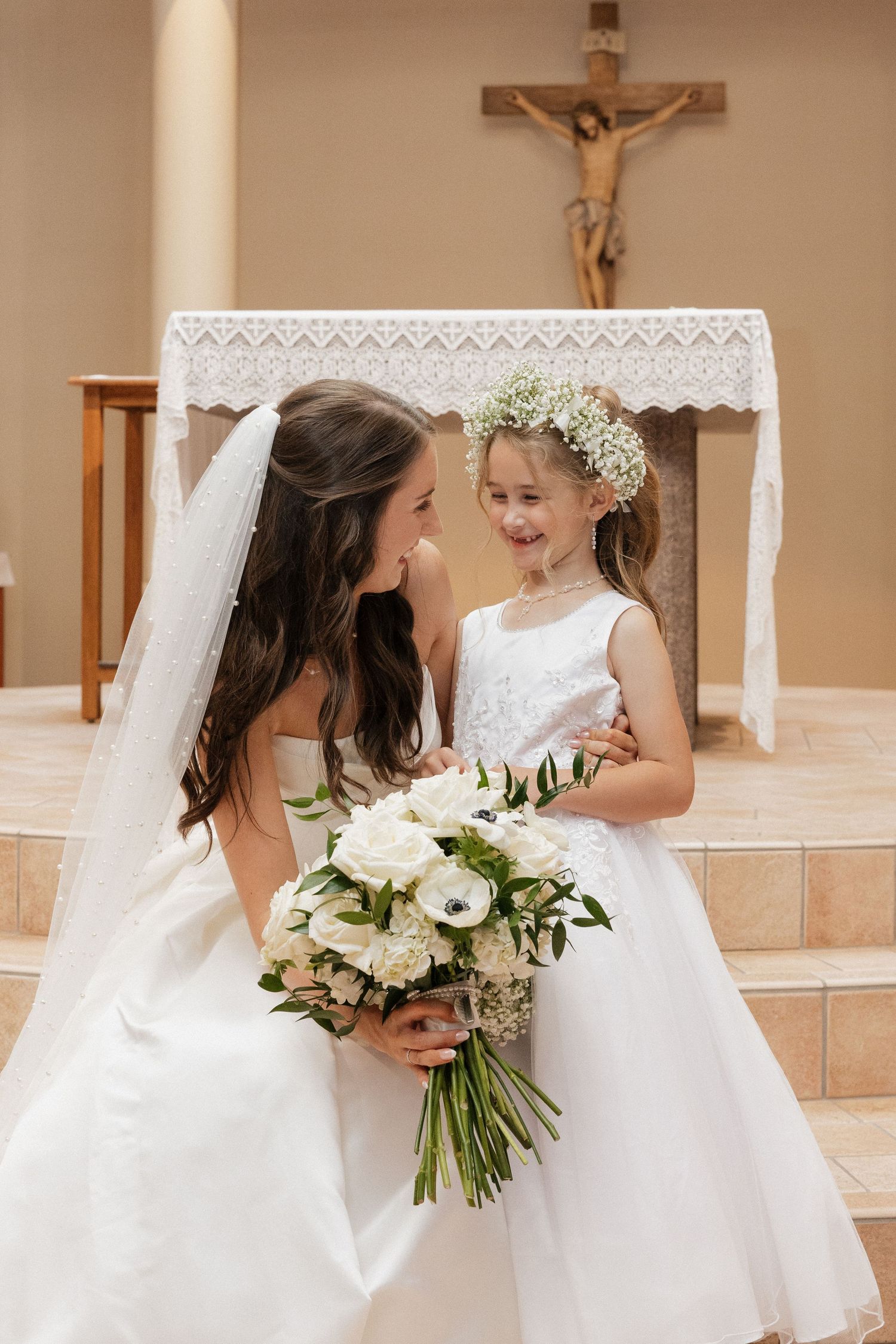 Bride in white gown holds bouquet beside flower girl in white dress with floral crown, smiling in a church.