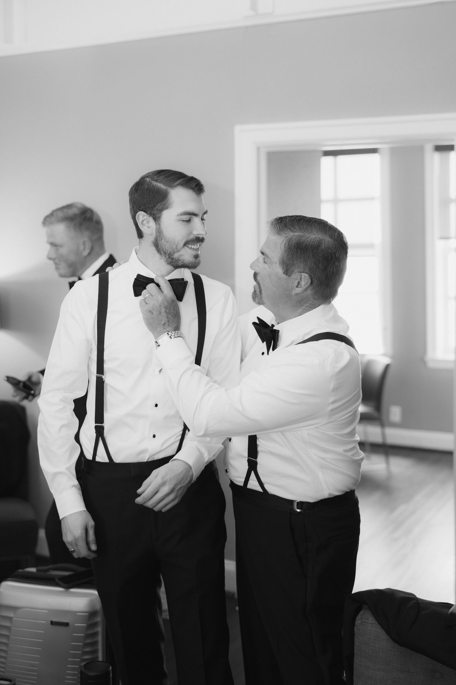 Two groomsmen in suspenders and bow ties help each other get dressed before a wedding ceremony.