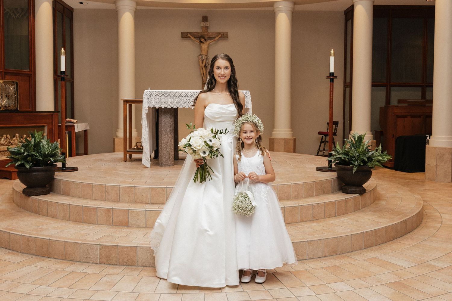 Bride in white gown with flower girl in white dress posing on church altar steps with bouquets.