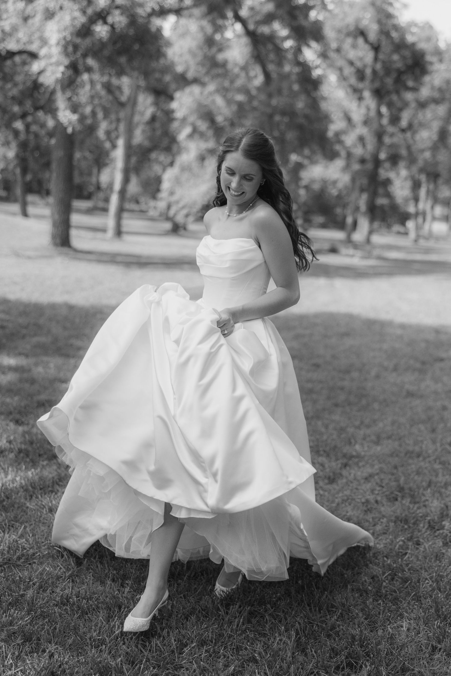 Black and white photo of a smiling bride in a strapless ball gown twirling in a lush green park.