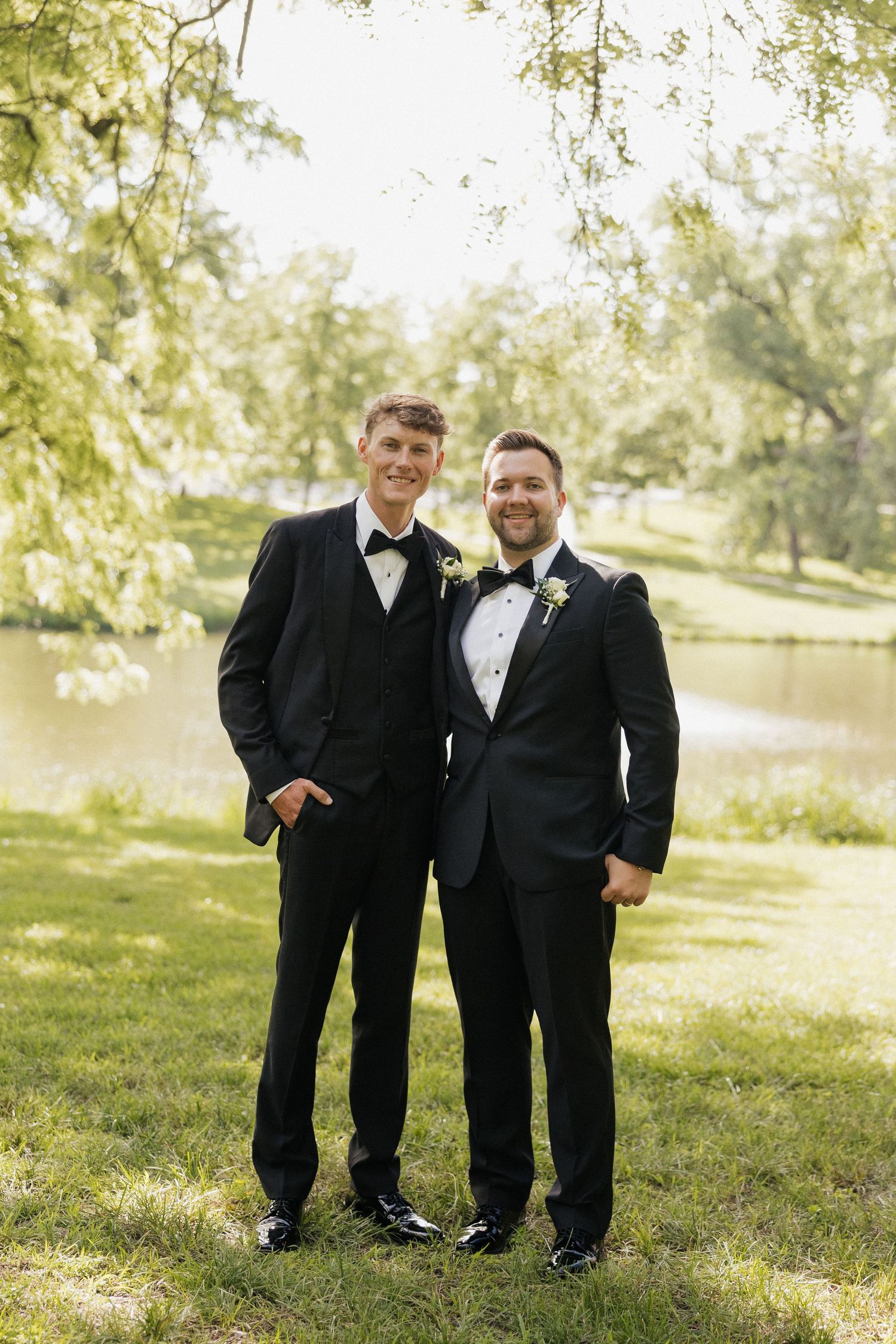 Two grooms in black tuxedos with boutonnieres smile outdoors near a pond on their wedding day.
