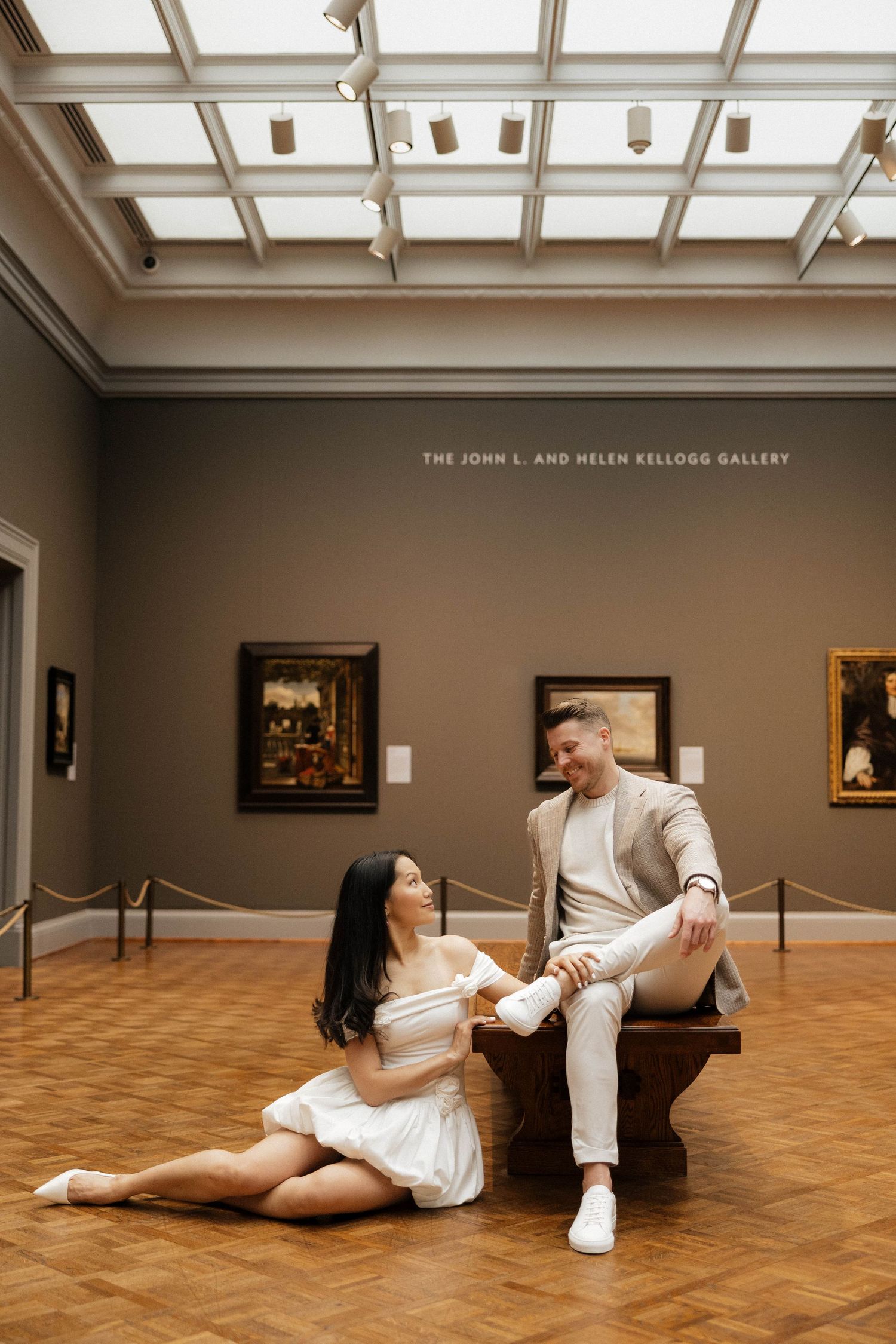 Couple posing in the John L. and Helen Kellogg Gallery, woman in white dress seated on floor, man in beige suit on bench.