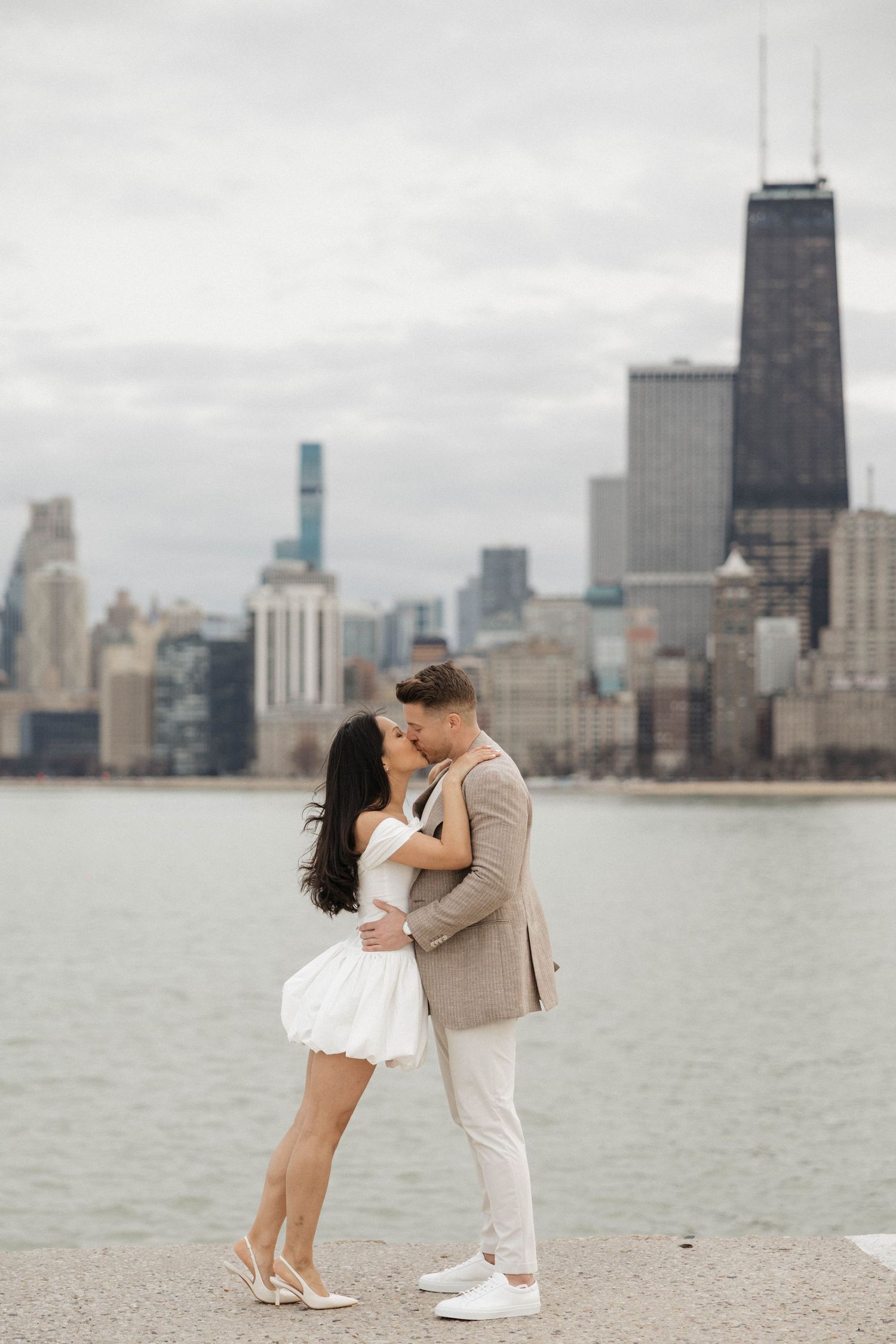 Couple sharing a romantic kiss by Lake Michigan with the Chicago skyline backdrop on a cloudy day.