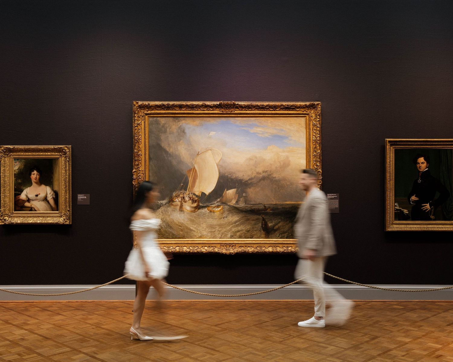 Two blurred visitors walk past a large Turner painting in a classic art museum gallery.