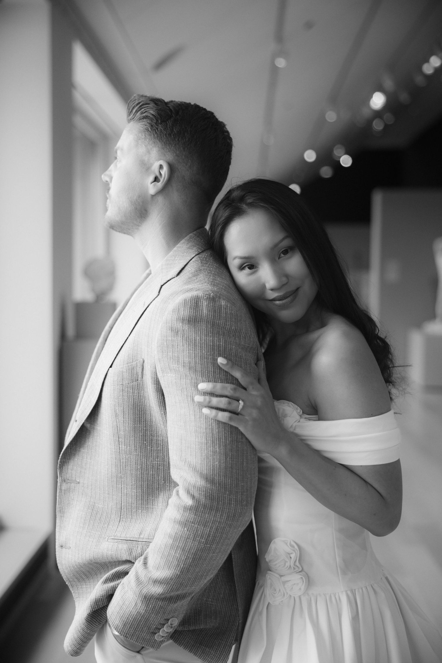 Black and white photo of a couple, woman in white off-shoulder dress leaning on mans back in suit jacket.