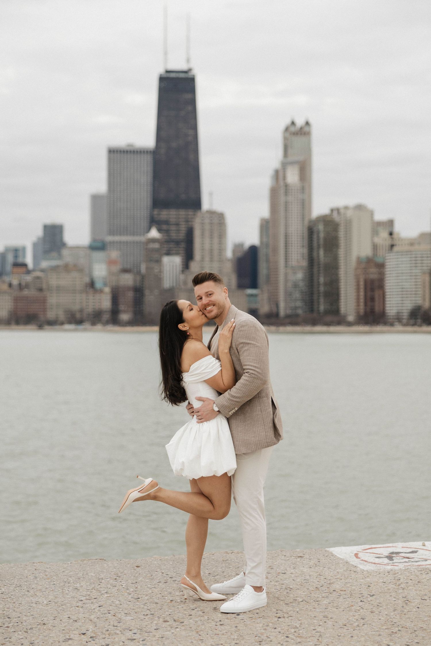 Couple embracing by Lake Michigan with Chicago skyline and John Hancock Center in background on cloudy day.