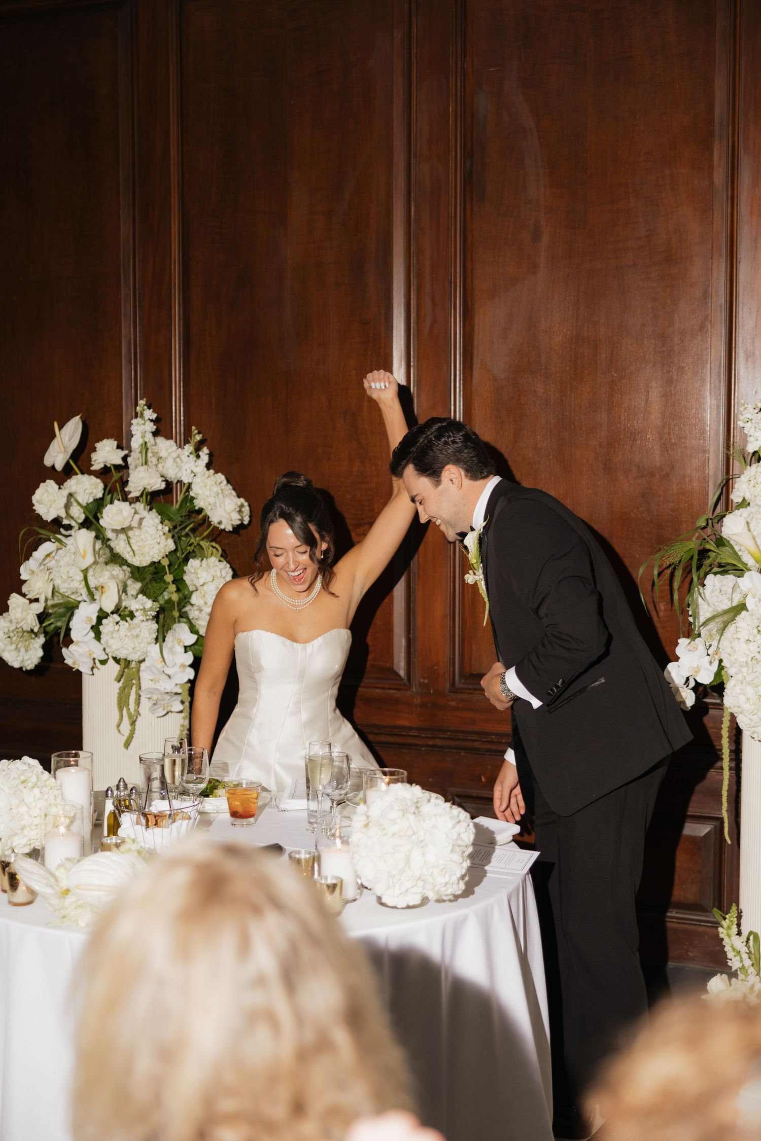 Bride and groom celebrating at wedding reception table with white floral arrangements in elegant wood-paneled venue.