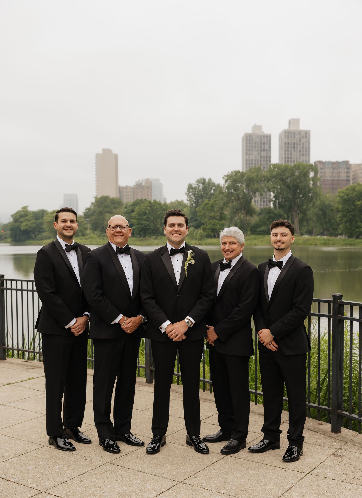 Five men in black tuxedos pose together outdoors near a lake with a city skyline backdrop on a wedding day.