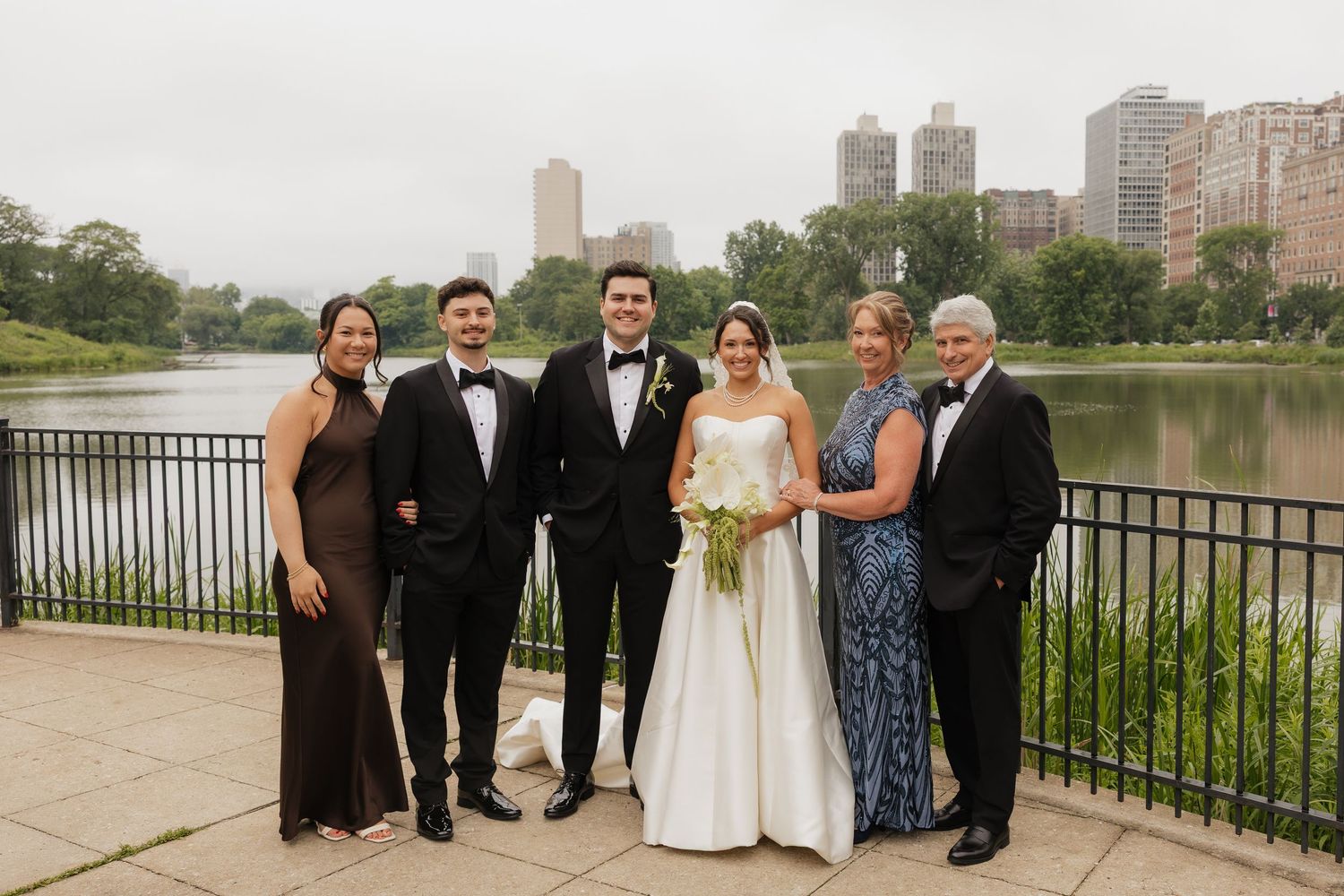 Wedding party of six poses by a lakeside railing with city skyline backdrop, bride in white gown holding bouquet.