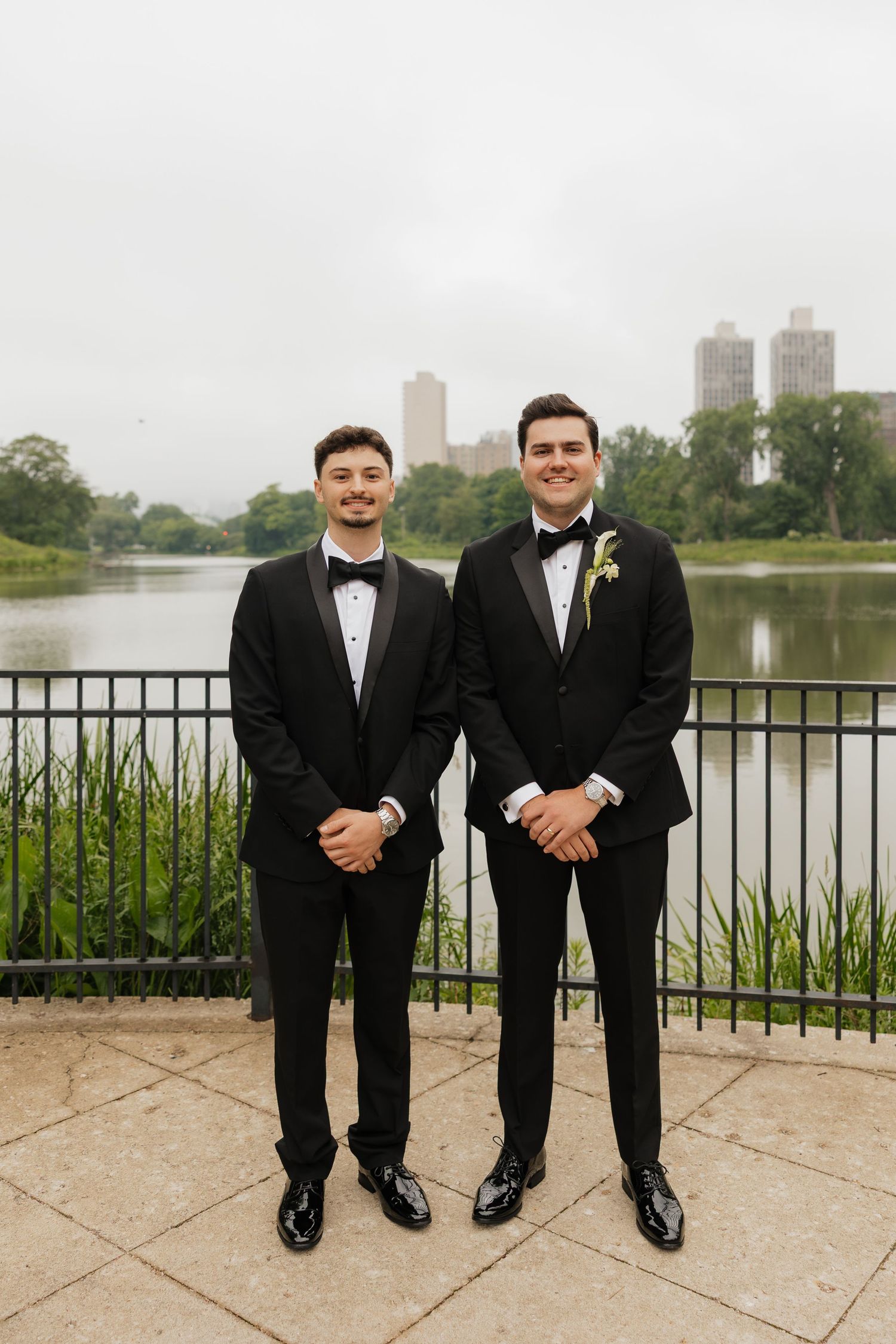 Two men in black tuxedos with bow ties pose outdoors near a lake with city skyline backdrop.