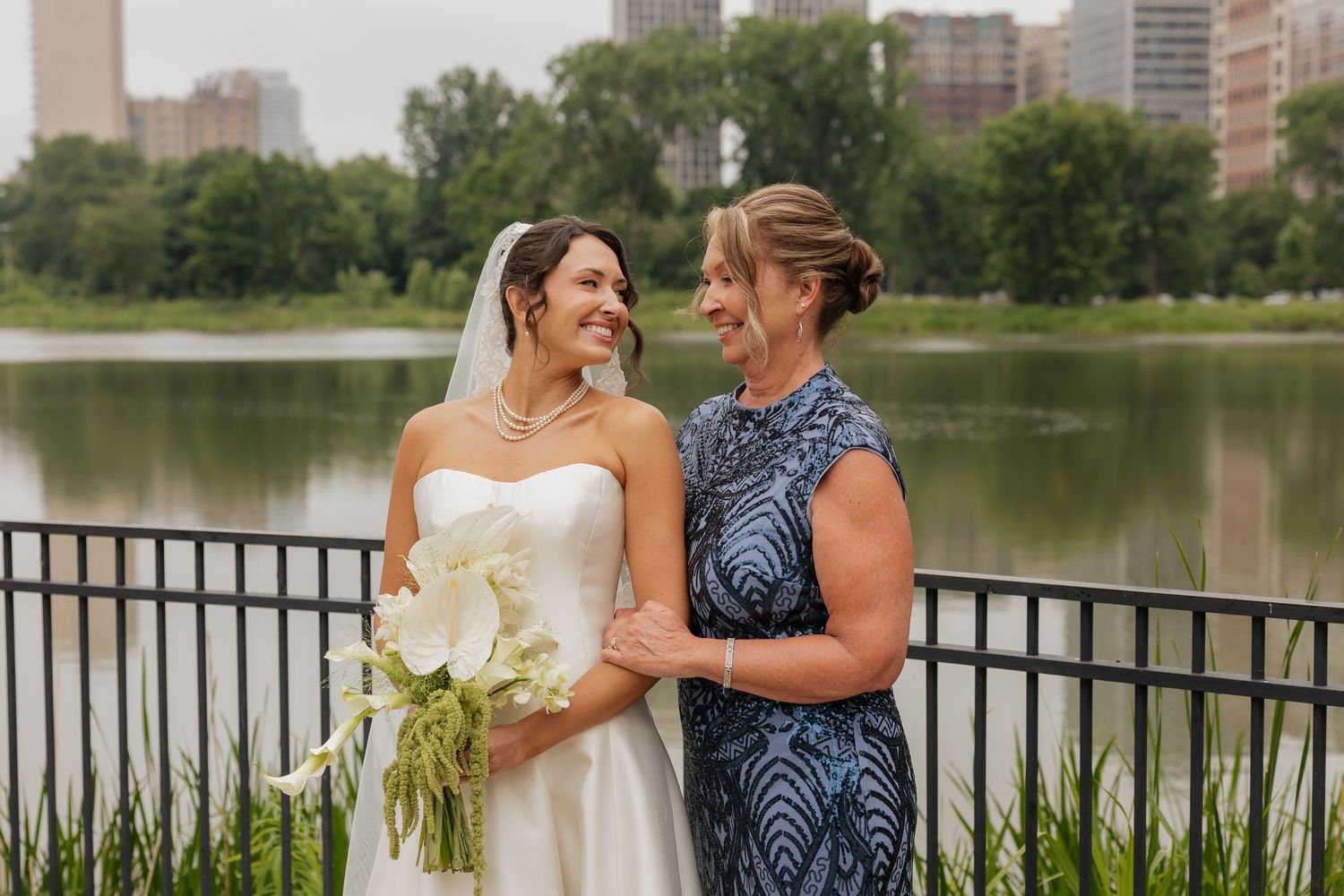 Bride in white gown holding calla lilies smiles with guest in blue dress by a lakeside railing in a city park.