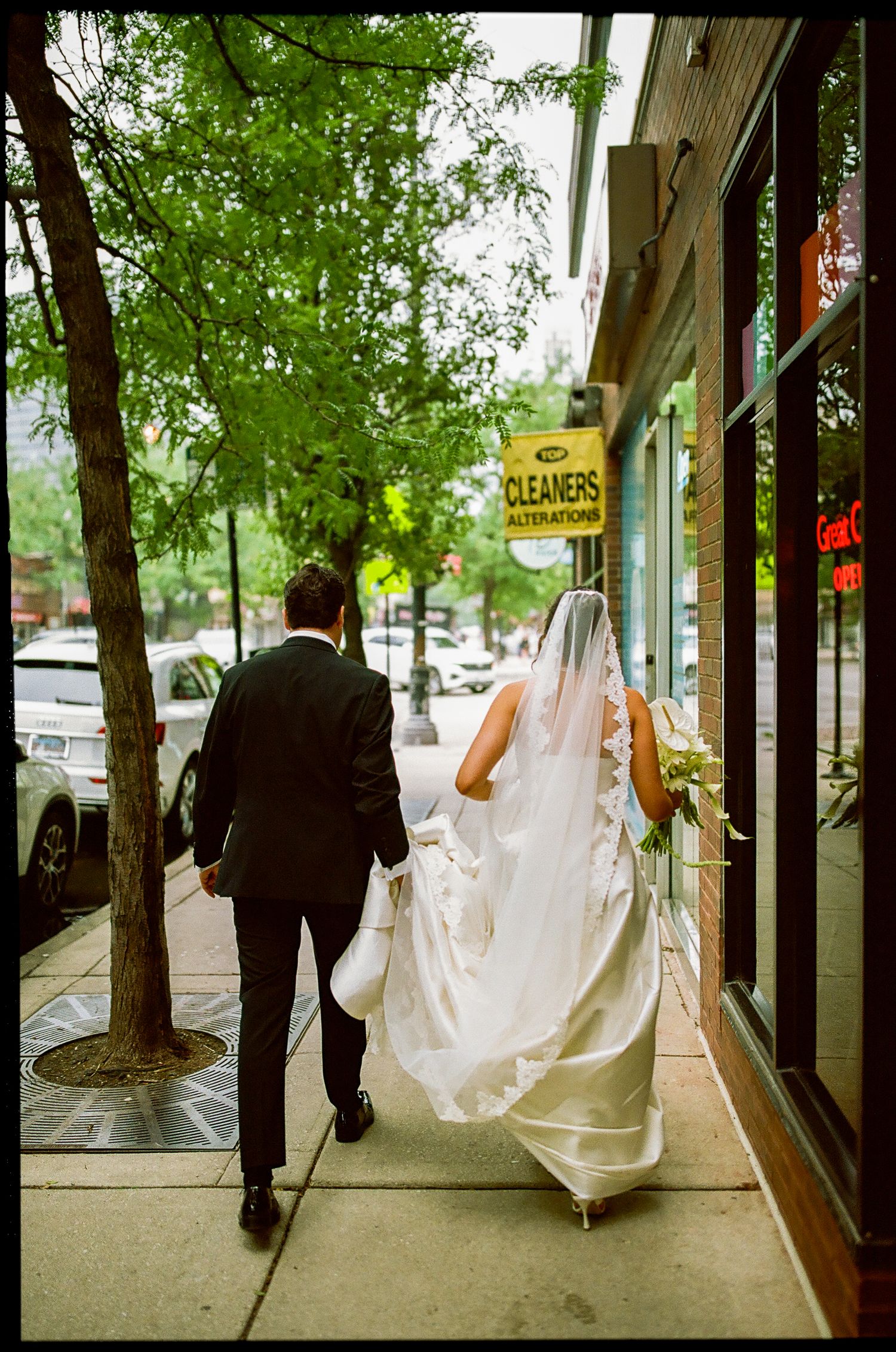 Bride and groom walking on city sidewalk, bride in white gown with veil, groom in black tuxedo.