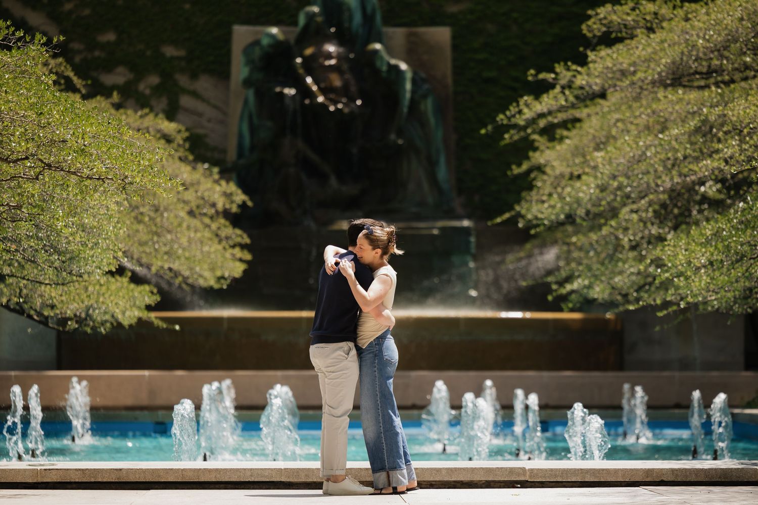 Couple embracing romantically in front of a fountain and bronze statue in a lush green park.