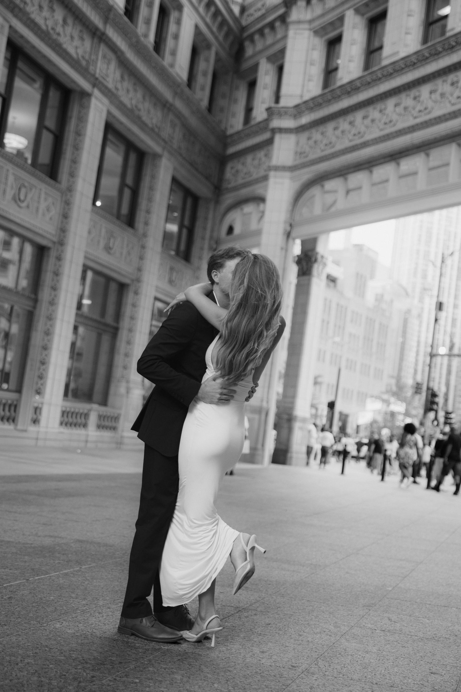 Black and white photo of a couple embracing and kissing in formal wear on a city street.