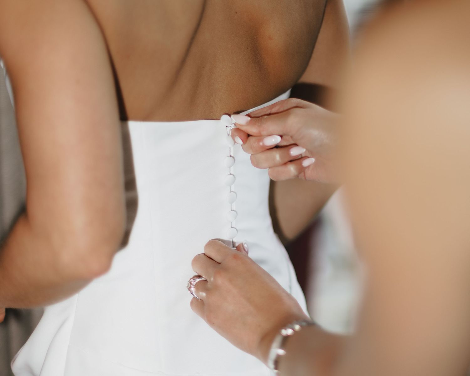 A bridesmaid carefully fastening delicate pearl buttons on the back of a white wedding gown.