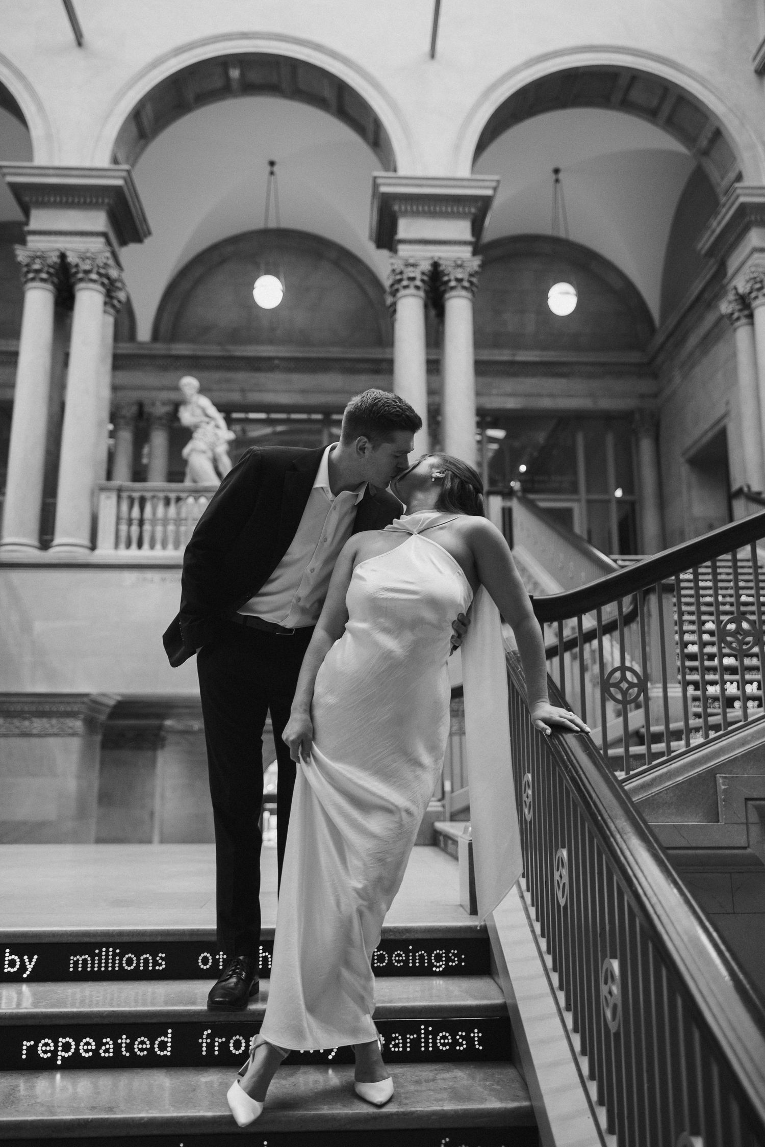 Black and white photo of a couple kissing on grand staircase in elegant venue, bride in white gown, groom in suit.