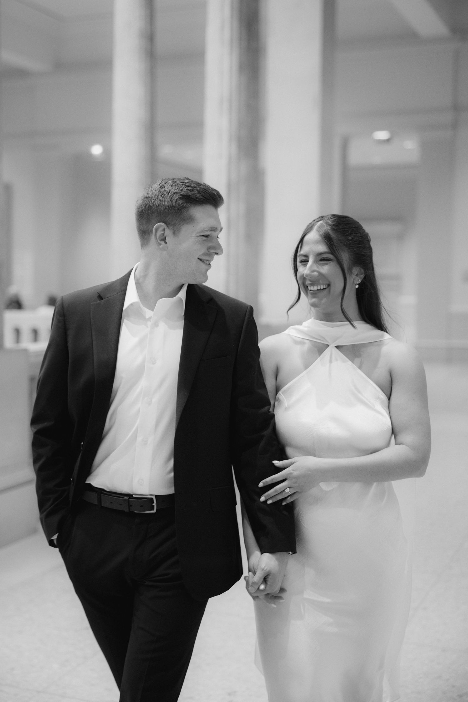 Black and white photo of a smiling couple in formal attire holding hands and walking together in an elegant venue.