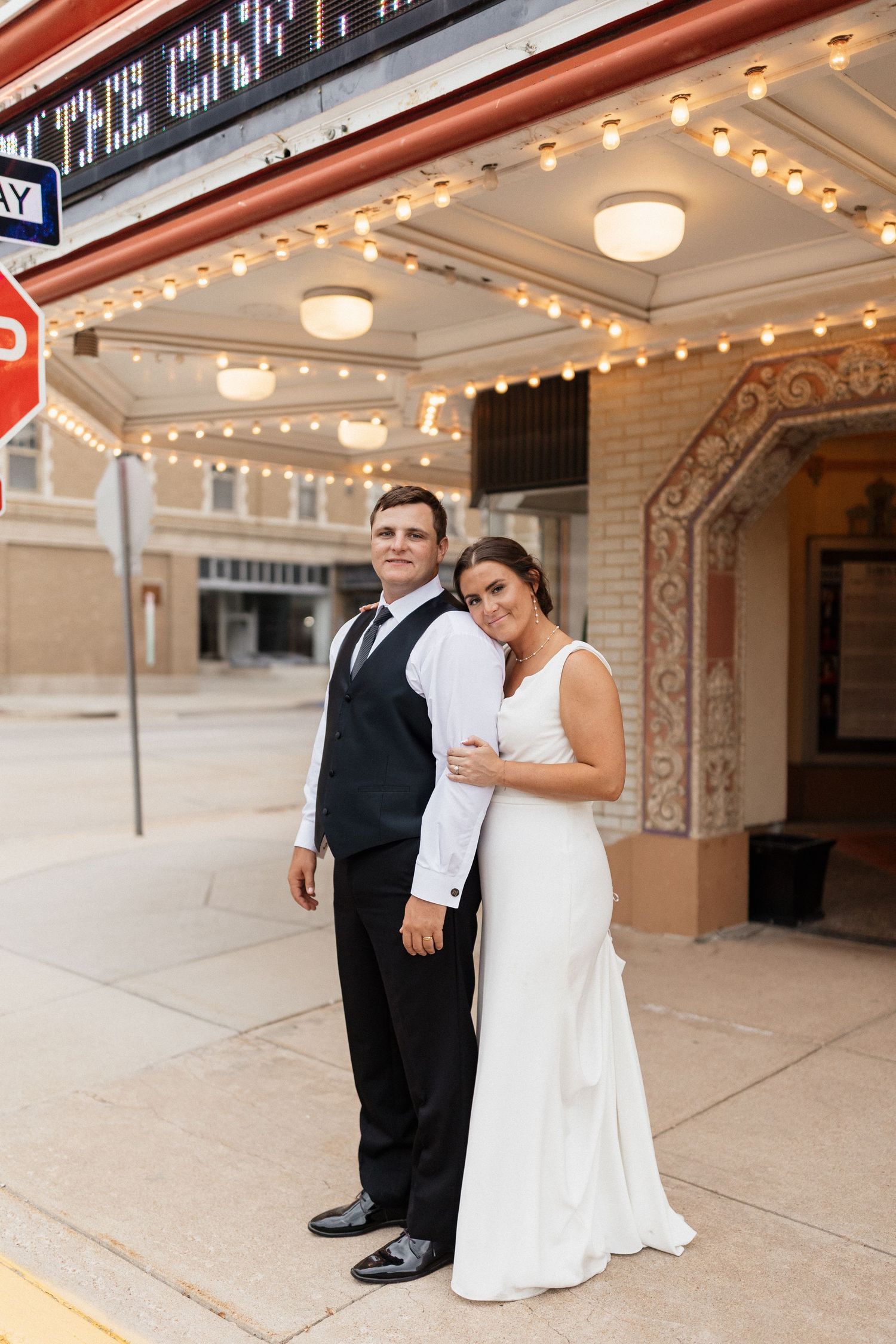 A newlywed couple poses outside a historic theater with ornate architecture and string lights.