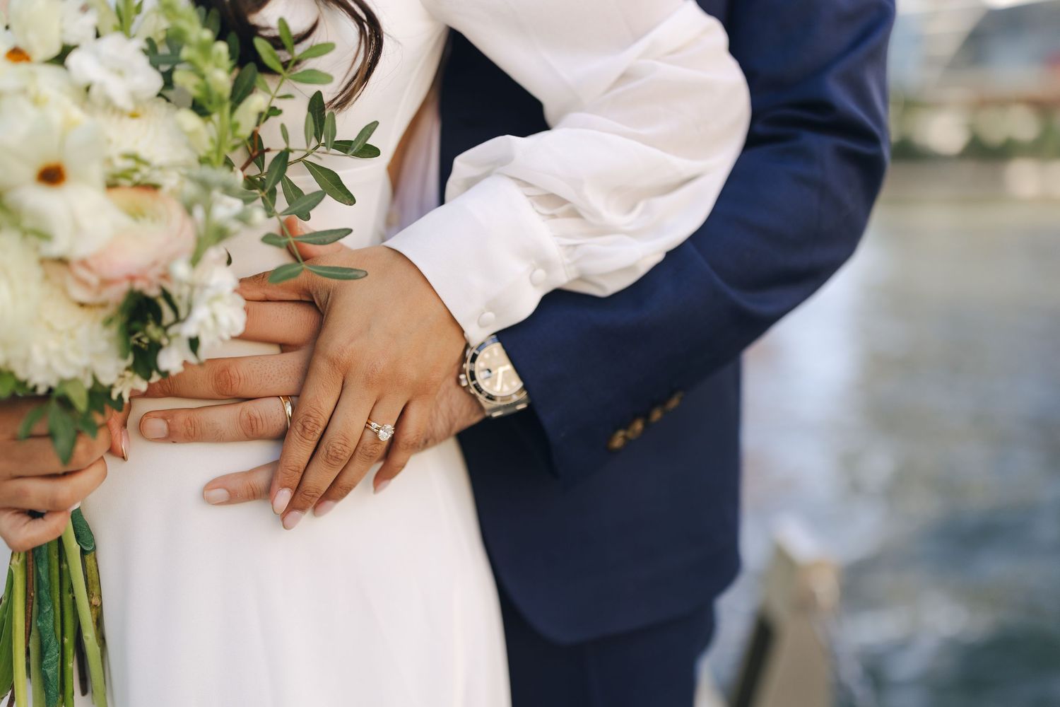 Close up of couple's hands holding white floral wedding bouquet with navy suit and white dress details.