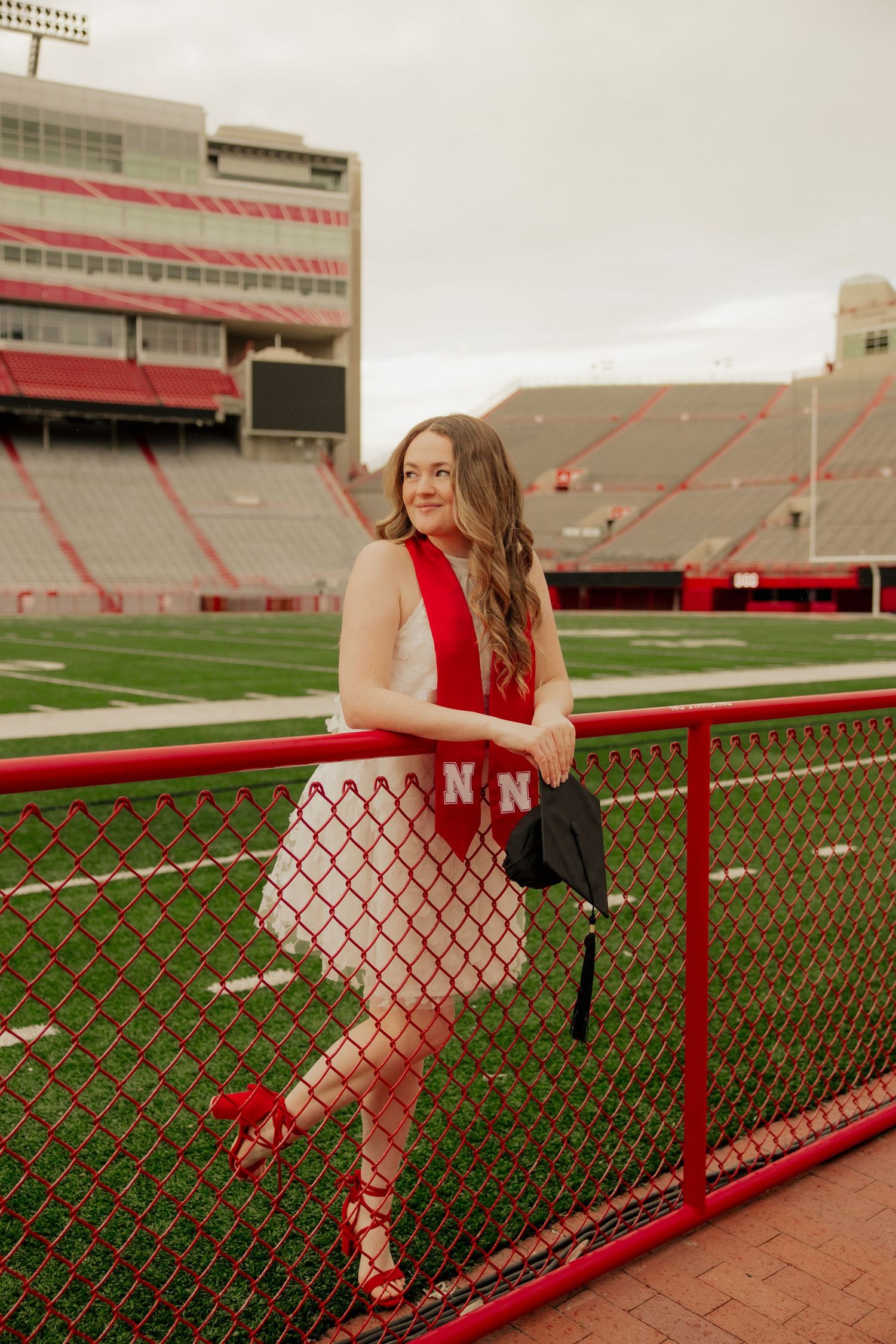 A person in a red dress poses on a railing at Memorial Stadium in Lincoln, Nebraska on a cloudy day.