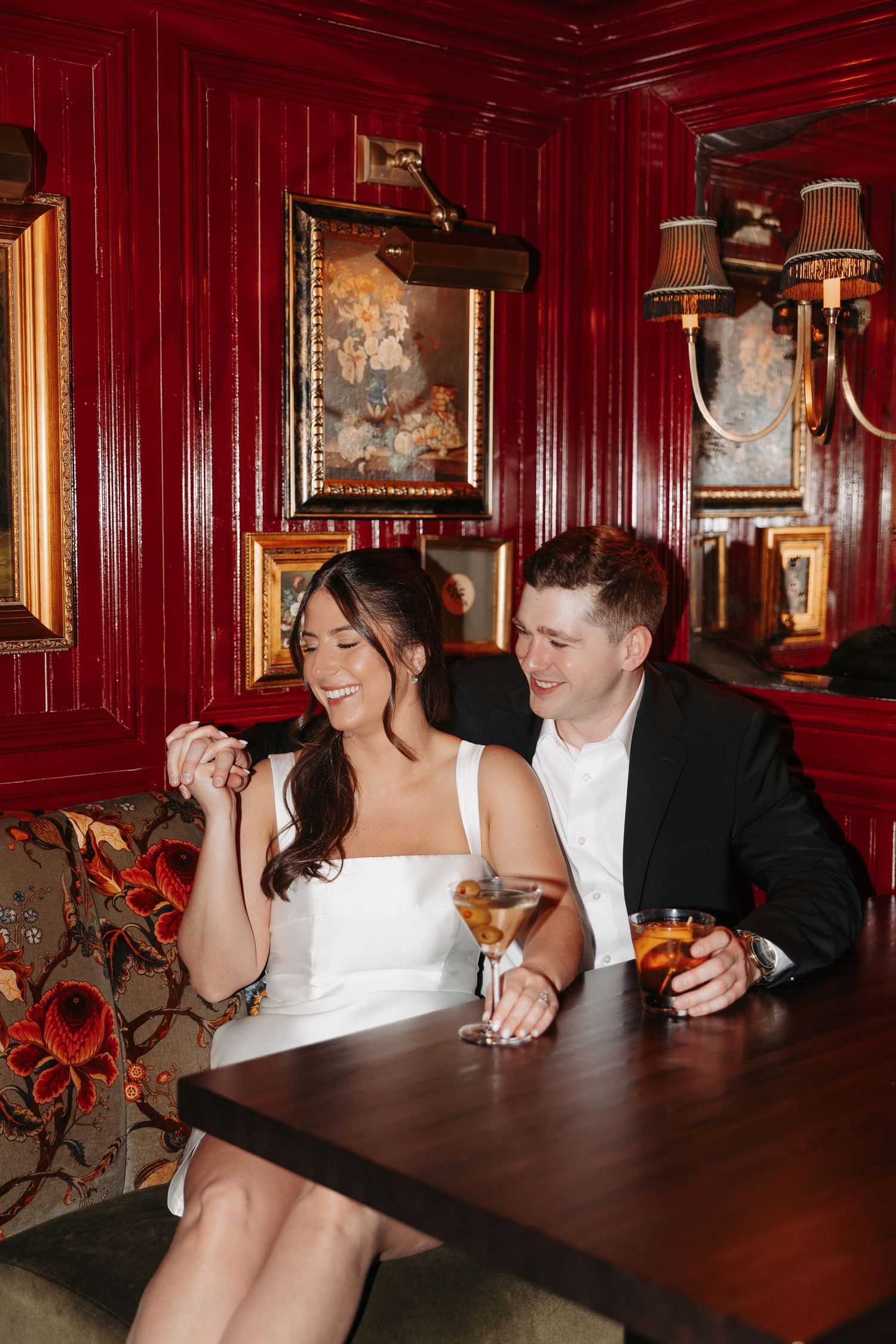 A couple in wedding attire shares a joyful moment at a table in an elegant red-walled restaurant interior.