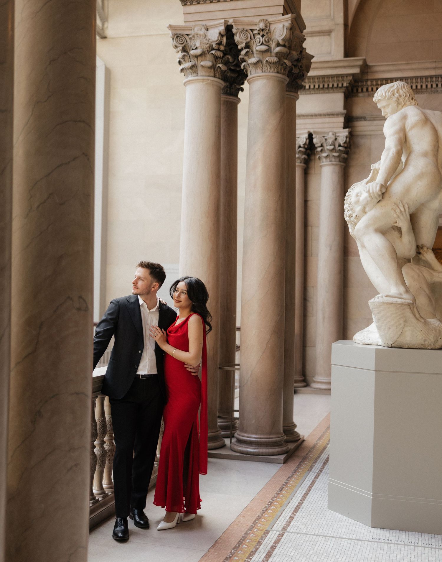 A couple in formal attire poses in an elegant museum gallery with classical columns and marble statues.