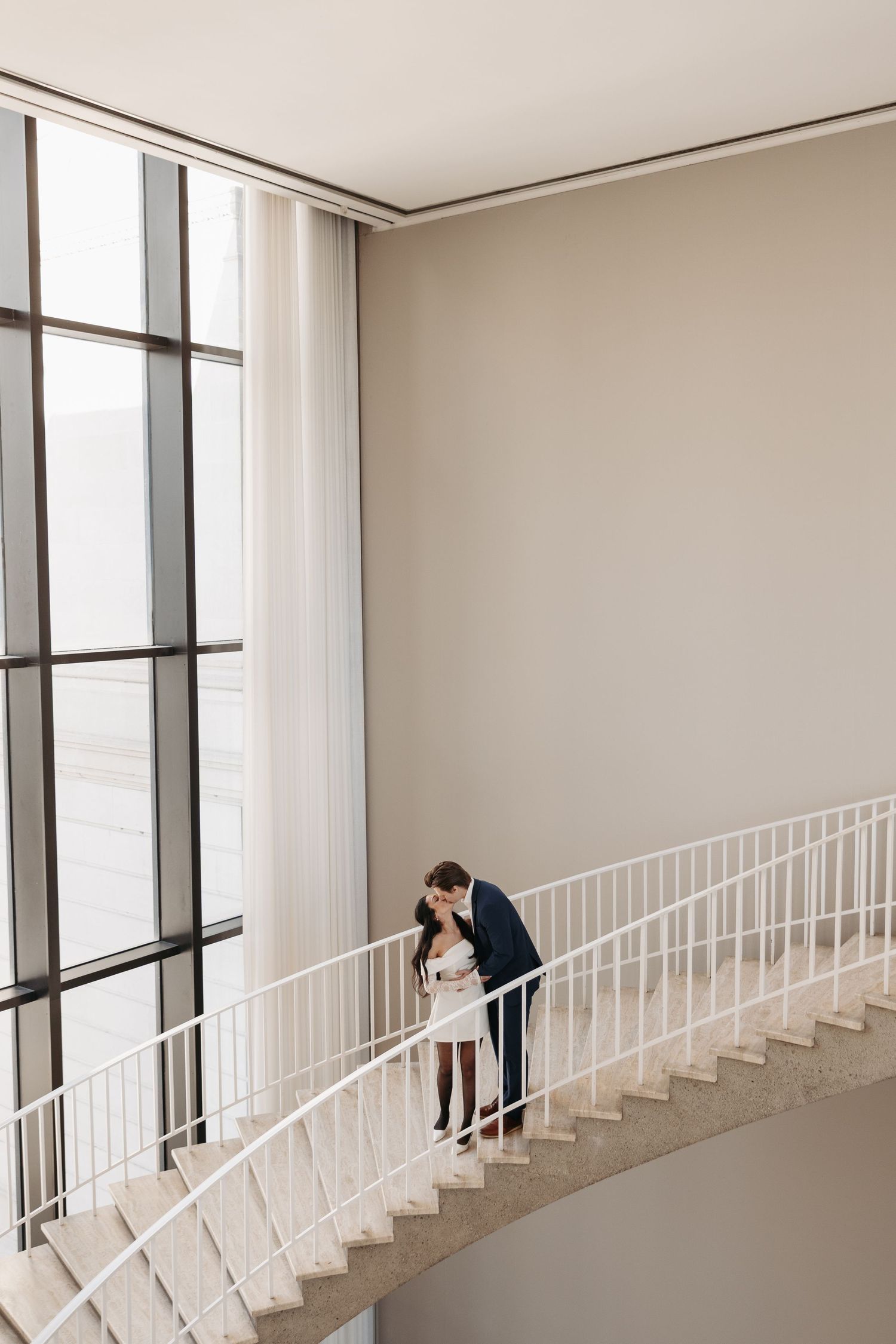 A couple shares a romantic moment on a curved white staircase with large windows.