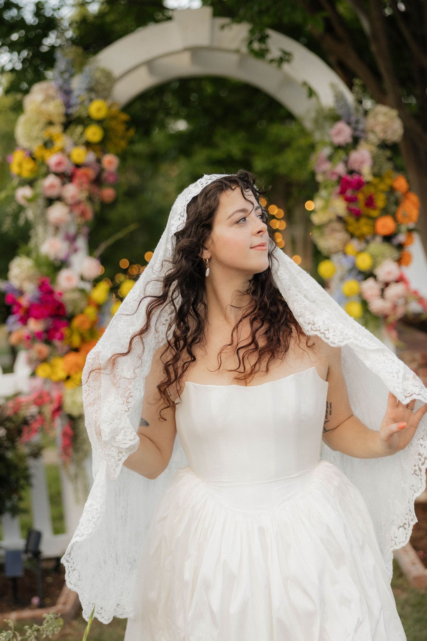 Beautiful bride in strapless white gown and lace veil posing before vibrant floral arch in outdoor garden setting.