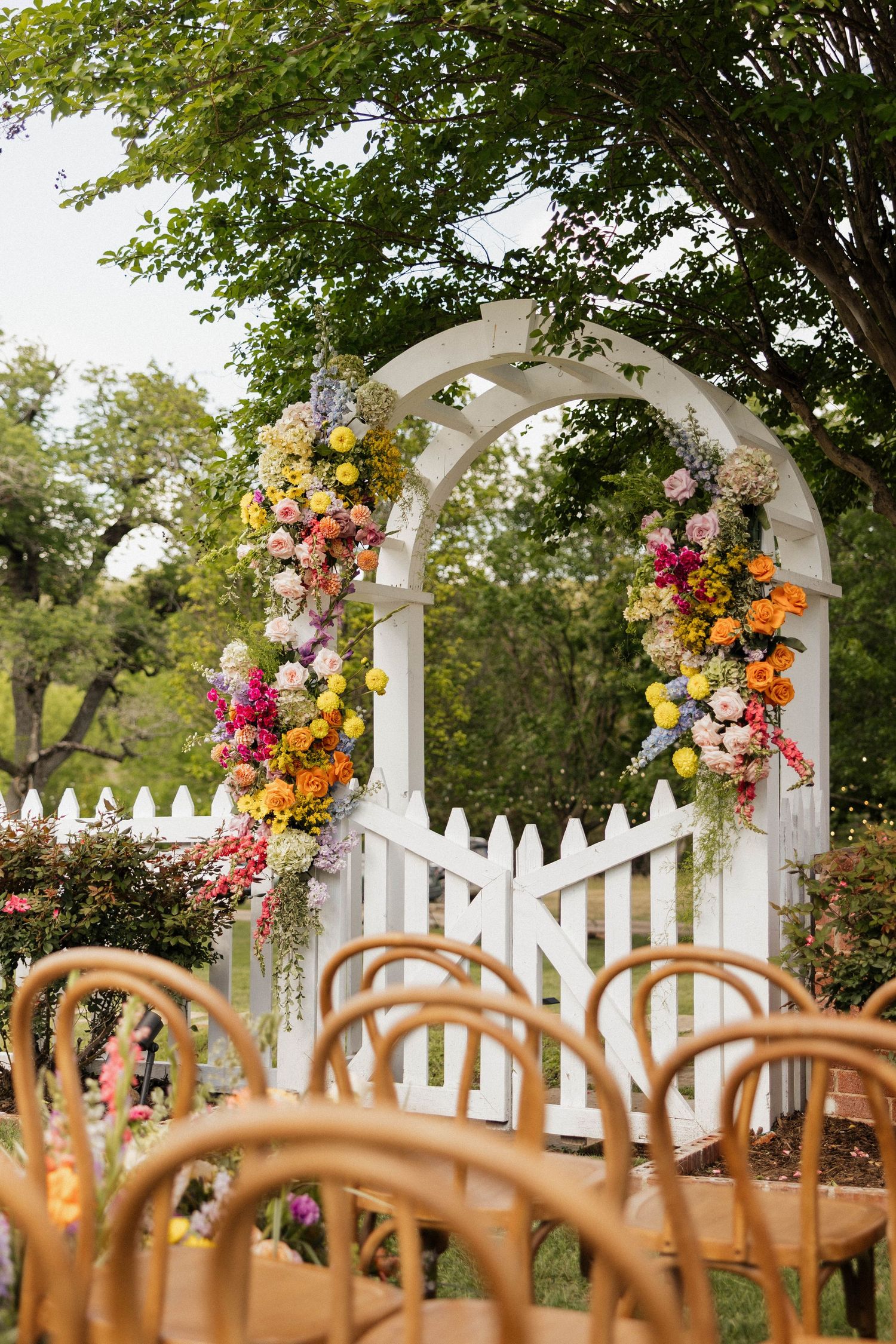 White garden arbor adorned with colorful floral wreaths at an outdoor wedding ceremony with wooden chairs.