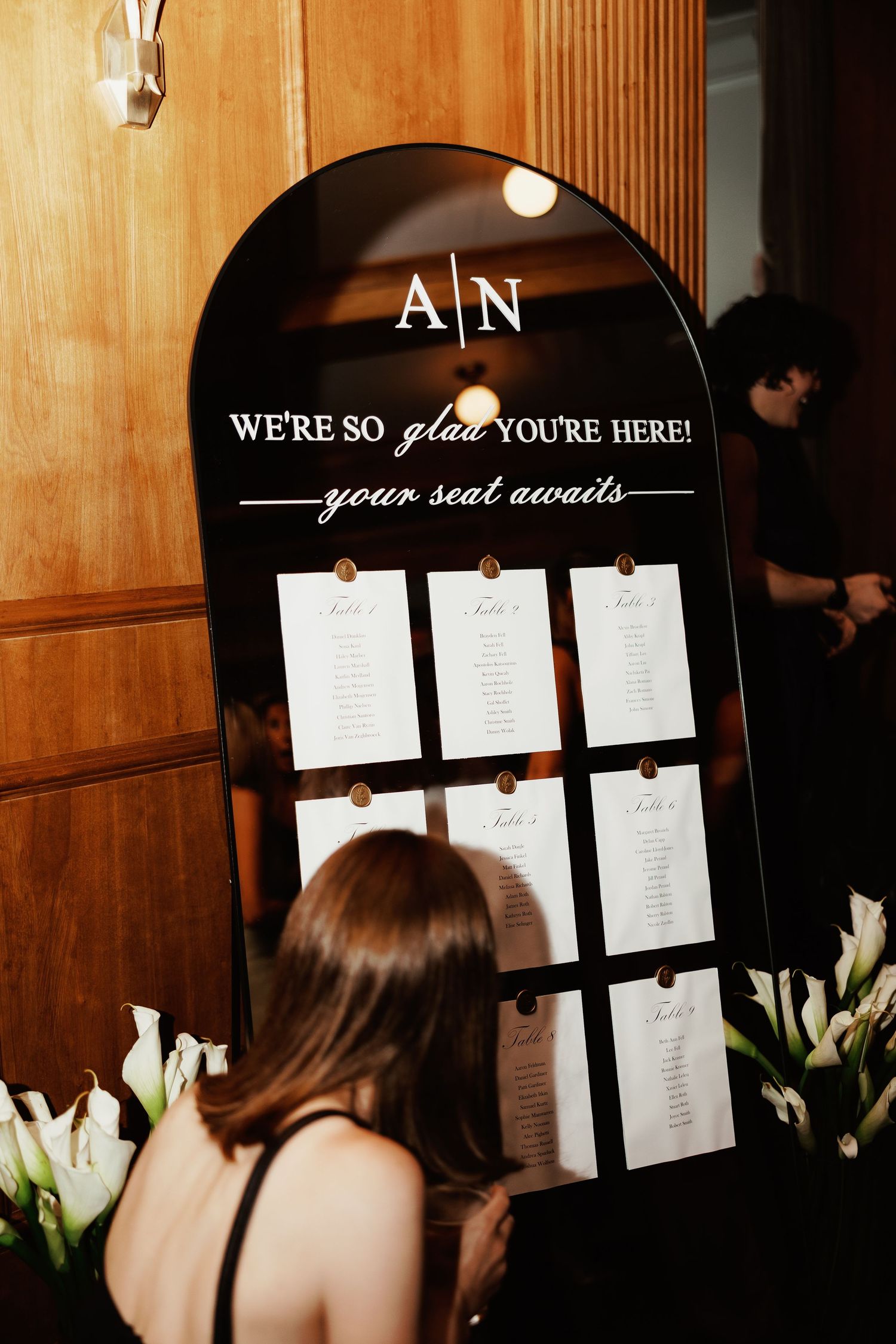 Black arched wedding seating chart display with white text reading We're So Glad You're Here, your seat awaits.