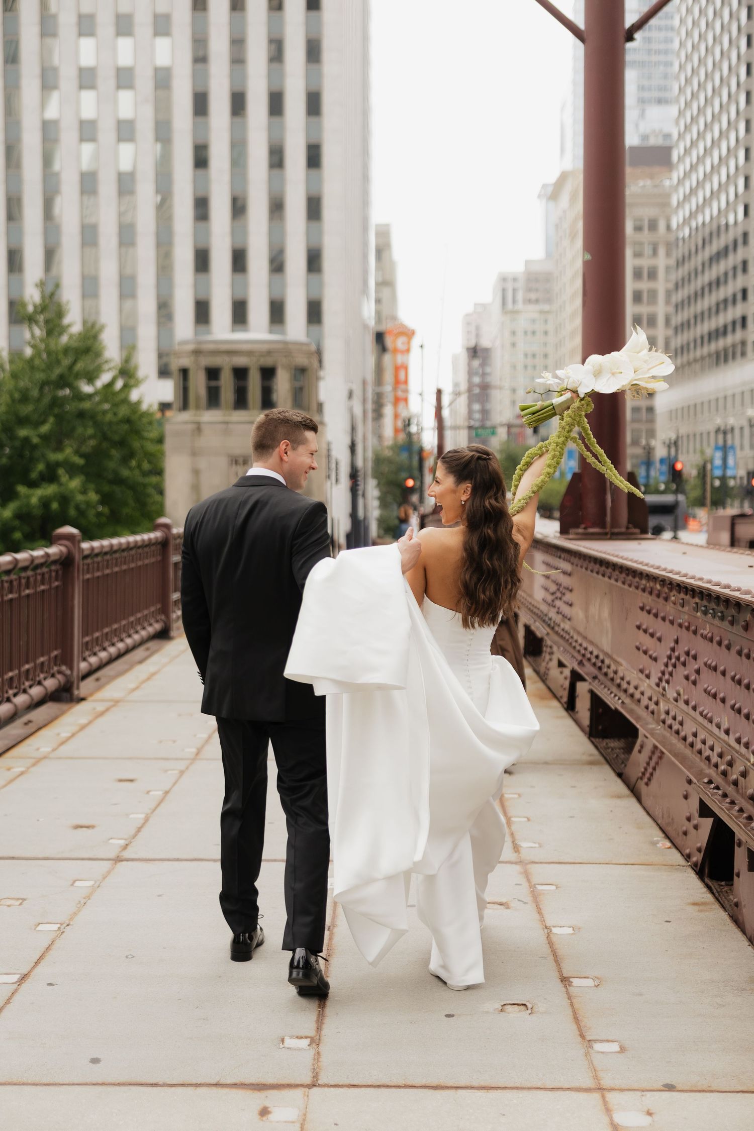 Bride and groom walking hand in hand on a city bridge, bride in white gown holding bouquet, groom in black tuxedo.