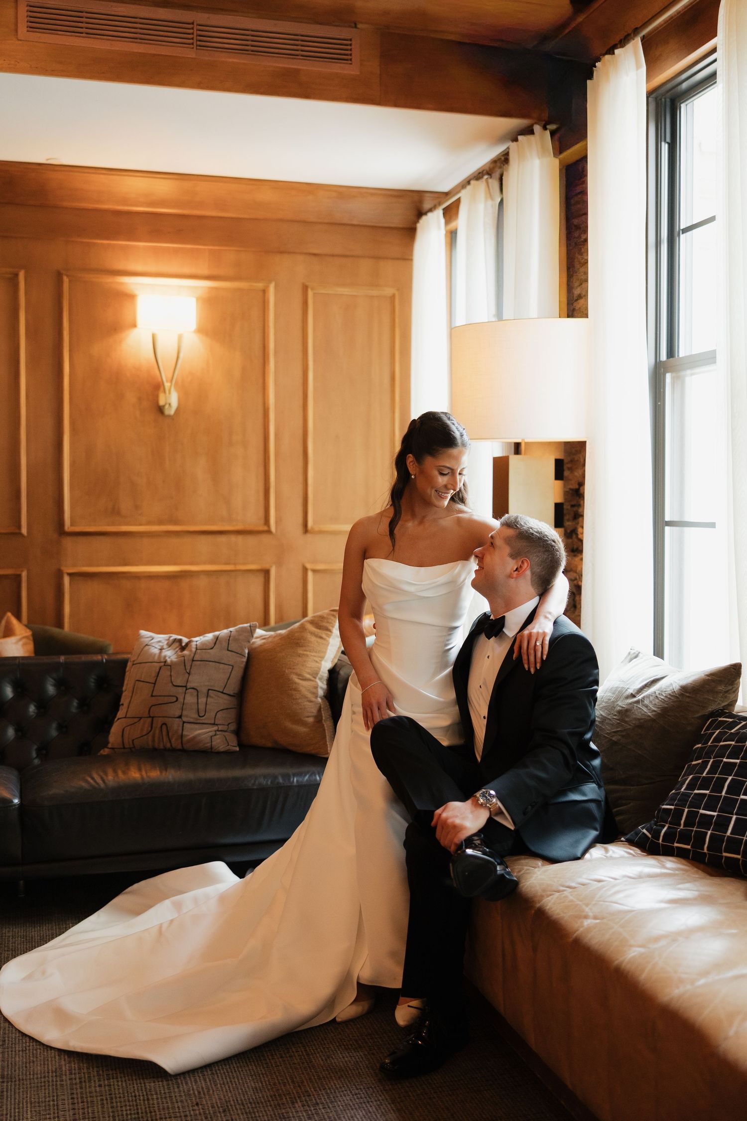 Bride in white gown and groom in black tuxedo share an intimate moment on a leather sofa in an elegant hotel suite.