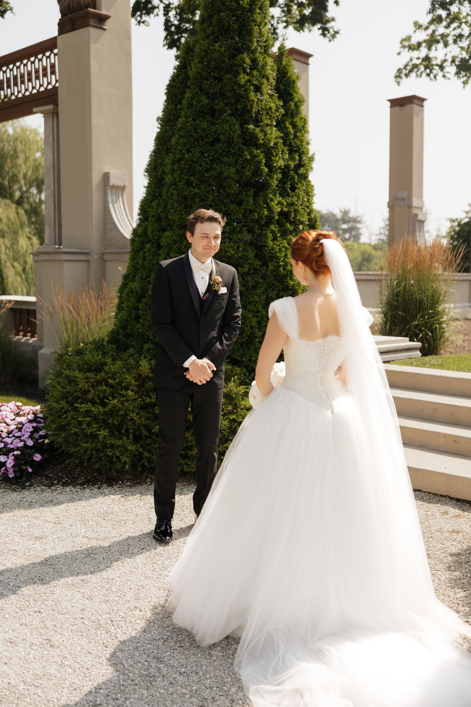 Groom in black suit smiles at bride in white ball gown during first look at elegant outdoor wedding venue.
