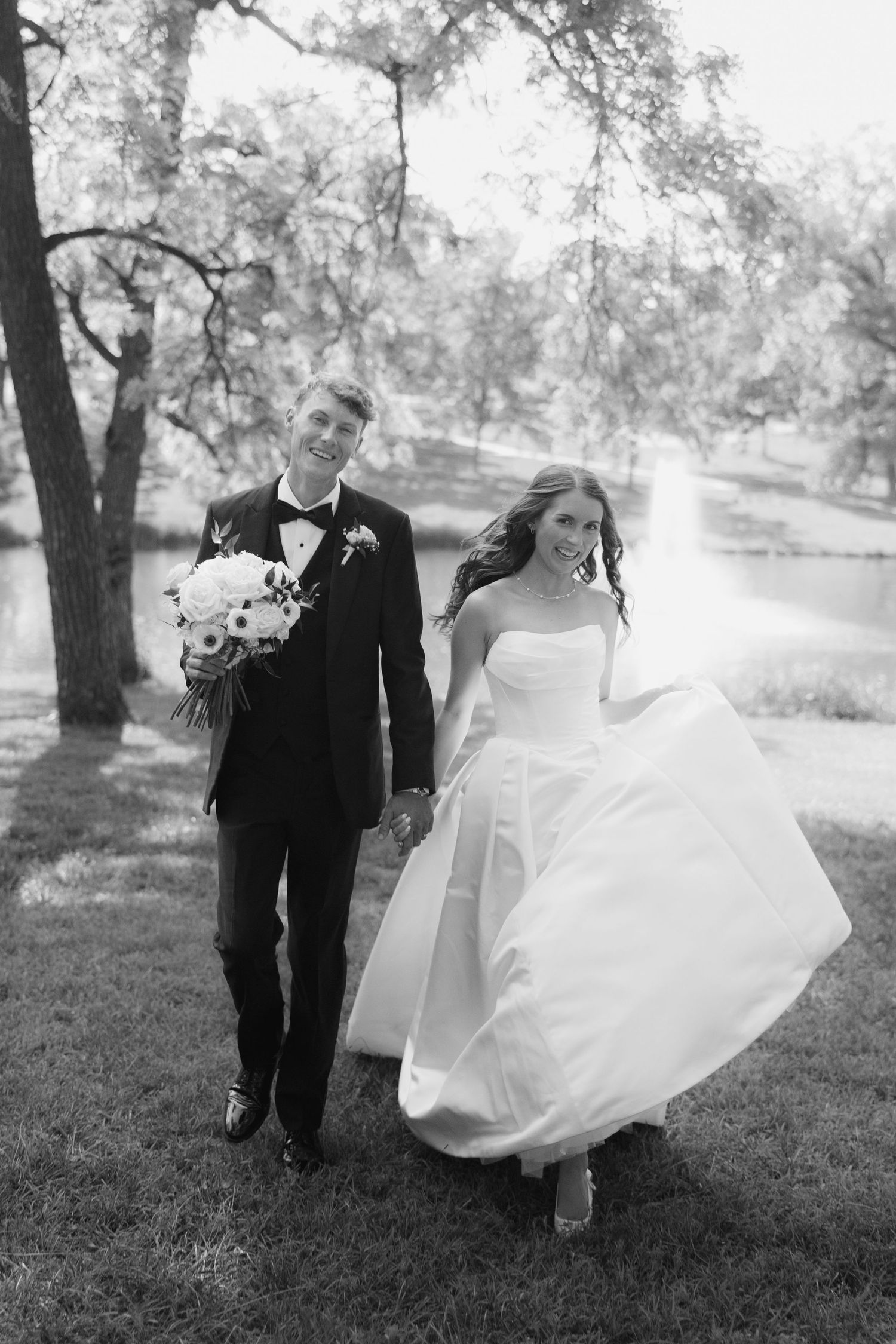 Black and white photo of smiling bride and groom walking hand in hand outdoors near trees and a pond.