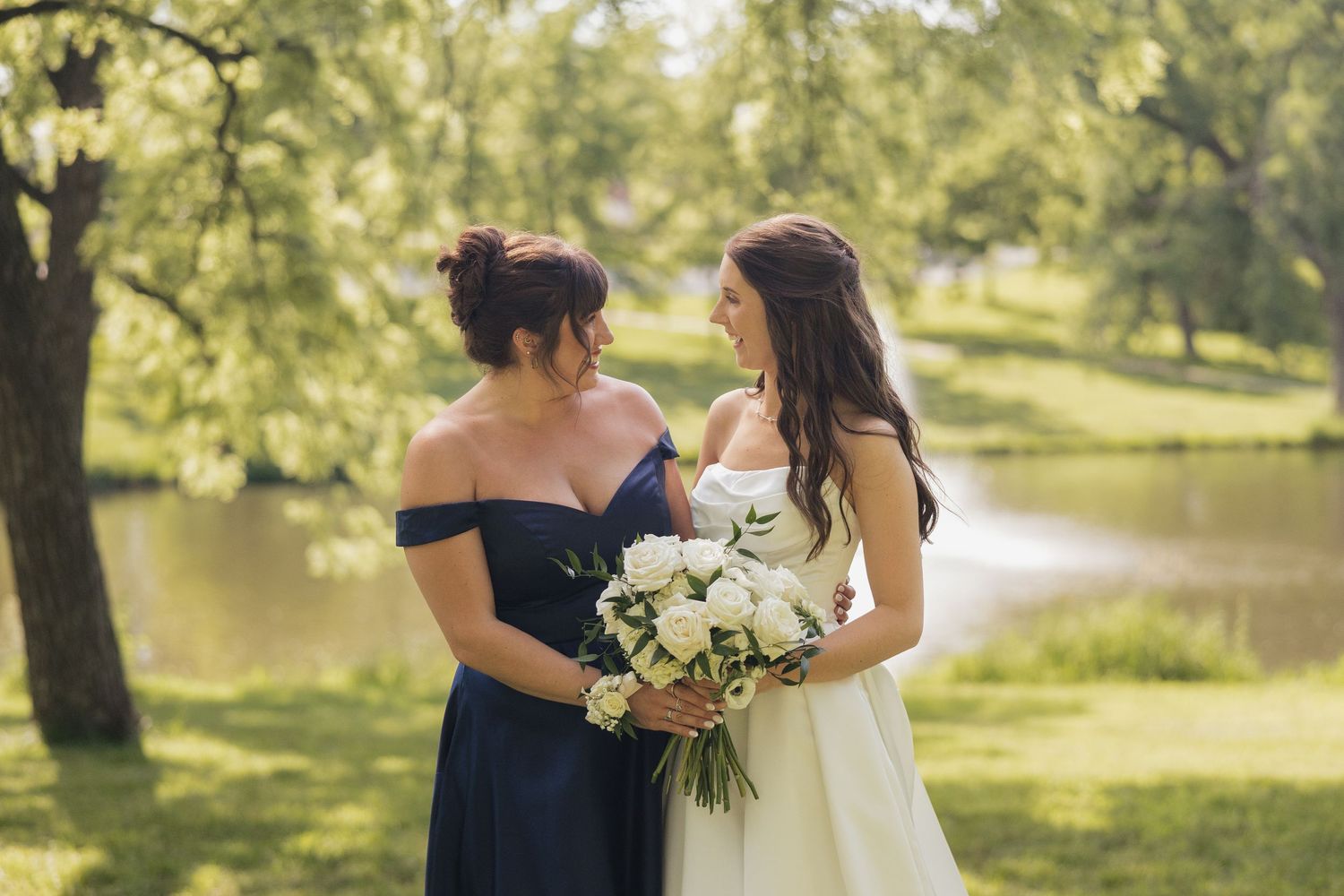Bride in white gown holds white bouquet beside bridesmaid in navy off-shoulder dress outdoors near pond.