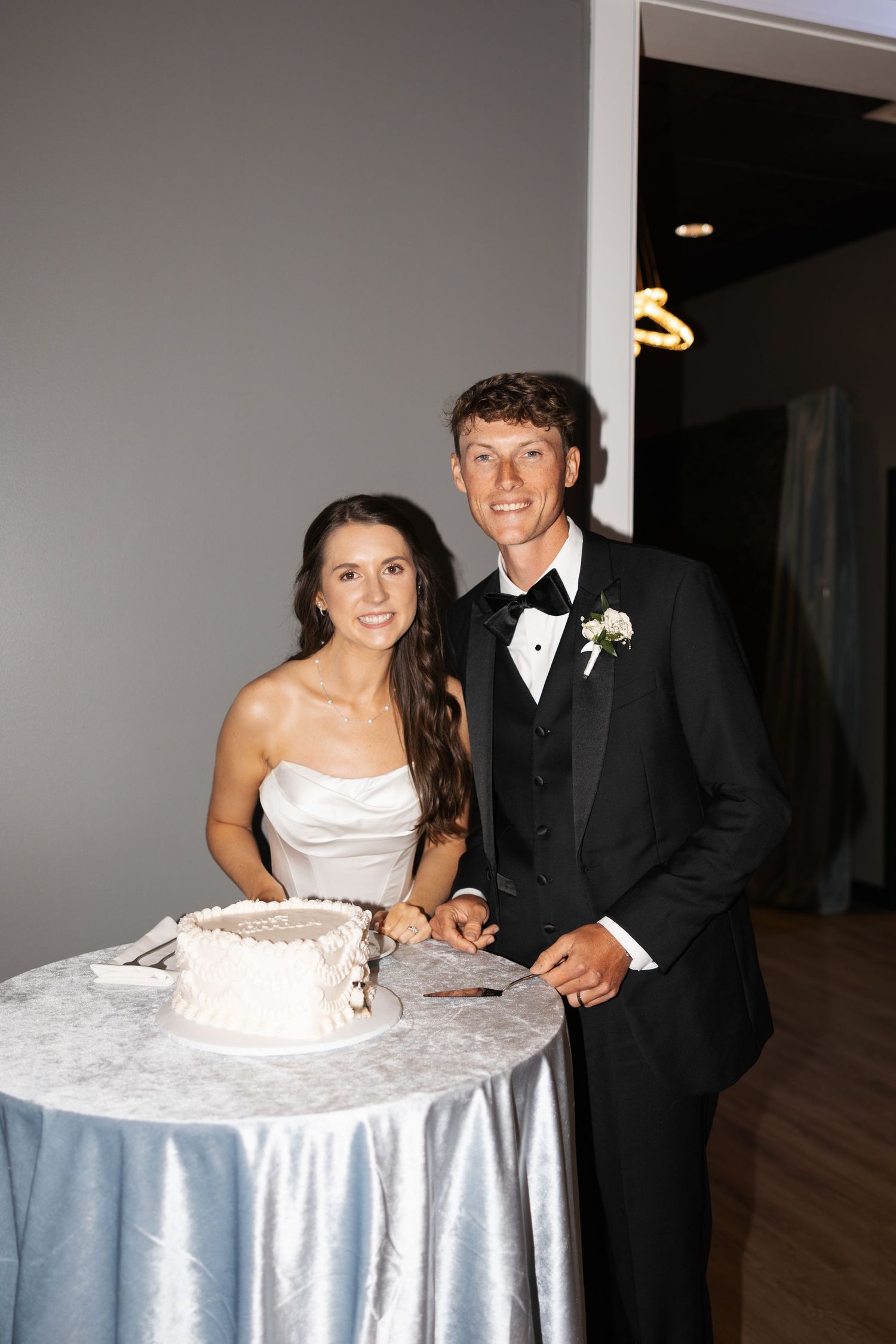 Smiling newlyweds in formal attire cutting their white wedding cake together at a reception venue.