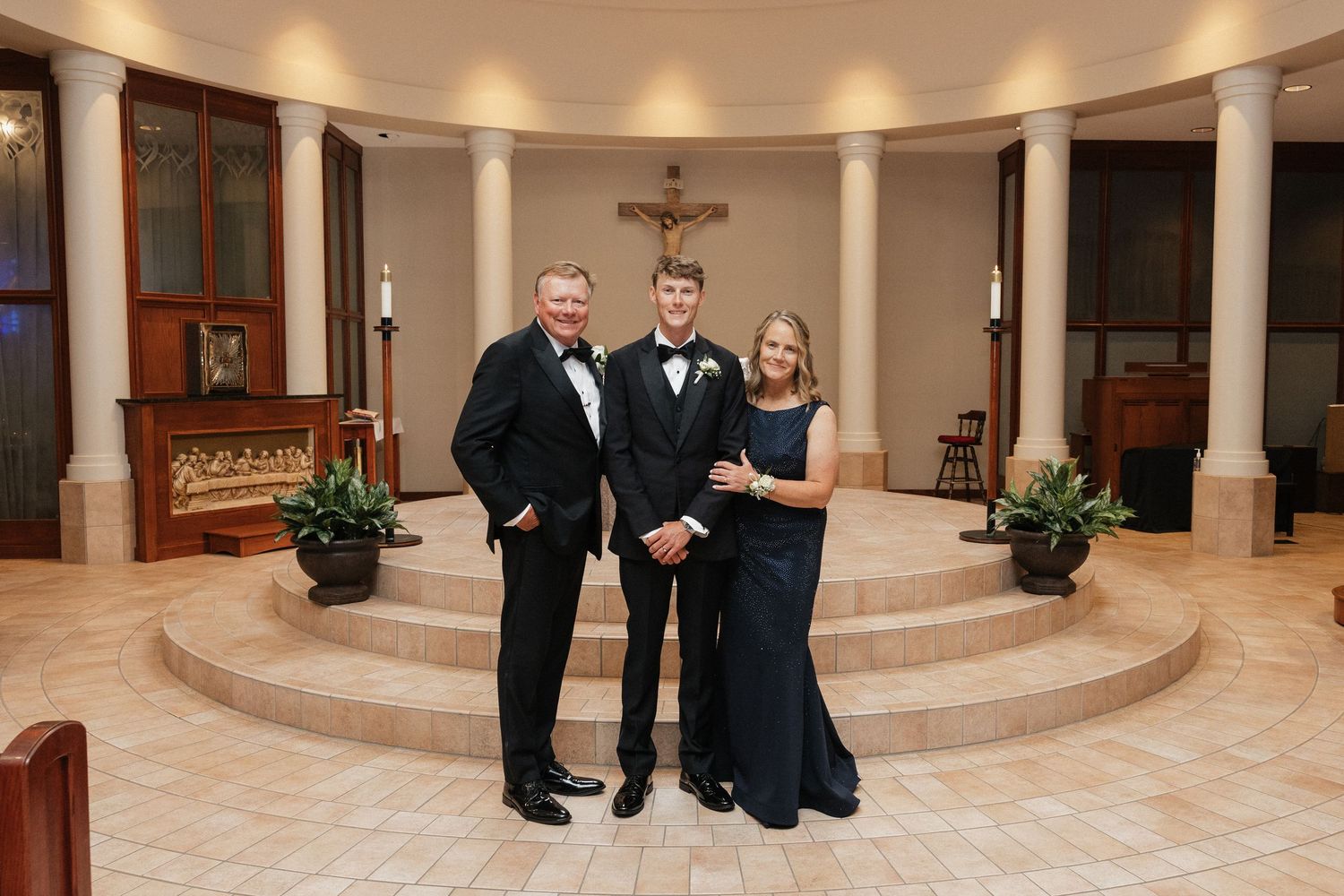 Family of three posing in formal attire inside an elegant chapel with a crucifix and columns.