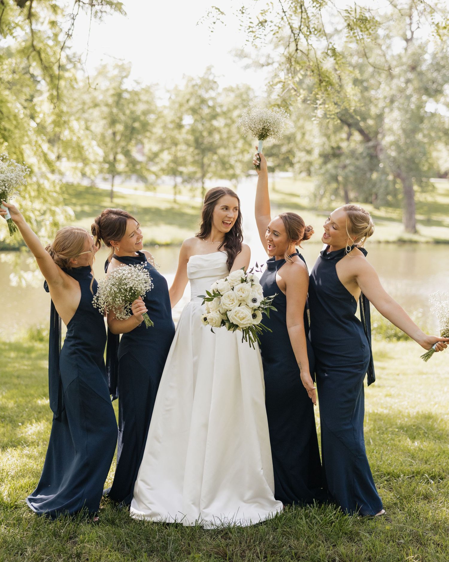 Bride in white gown with four bridesmaids in navy dresses celebrating outdoors near a pond with bouquets raised.