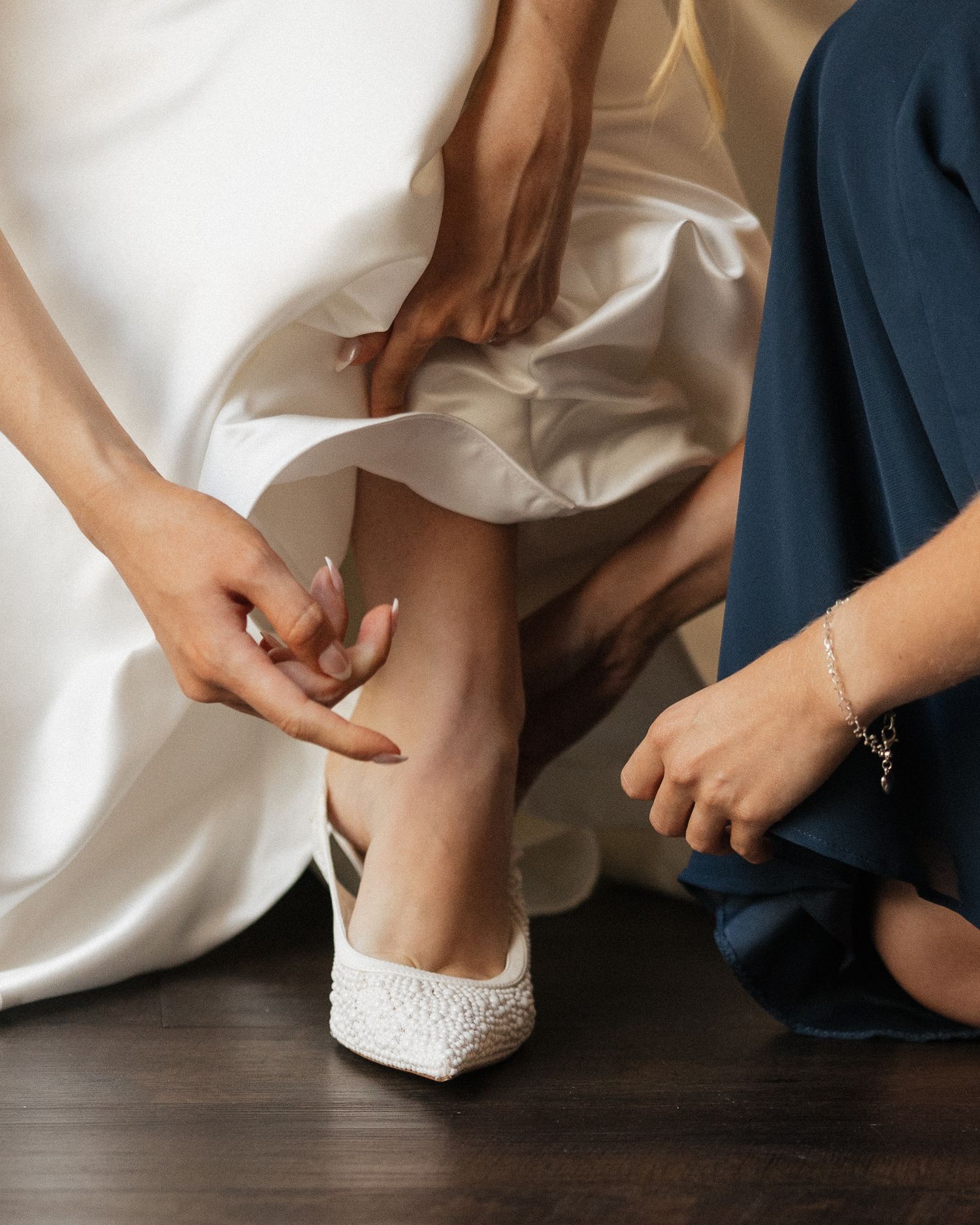 Bride in white gown having pearl wedding shoes put on by bridesmaid in blue dress.