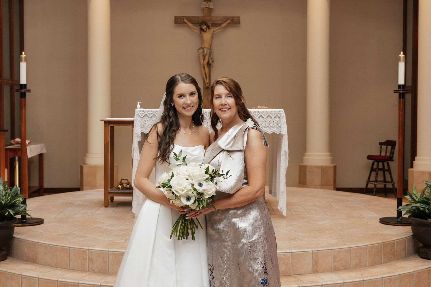 Bride in white gown holding bouquet stands with woman in silver dress before church altar with crucifix.