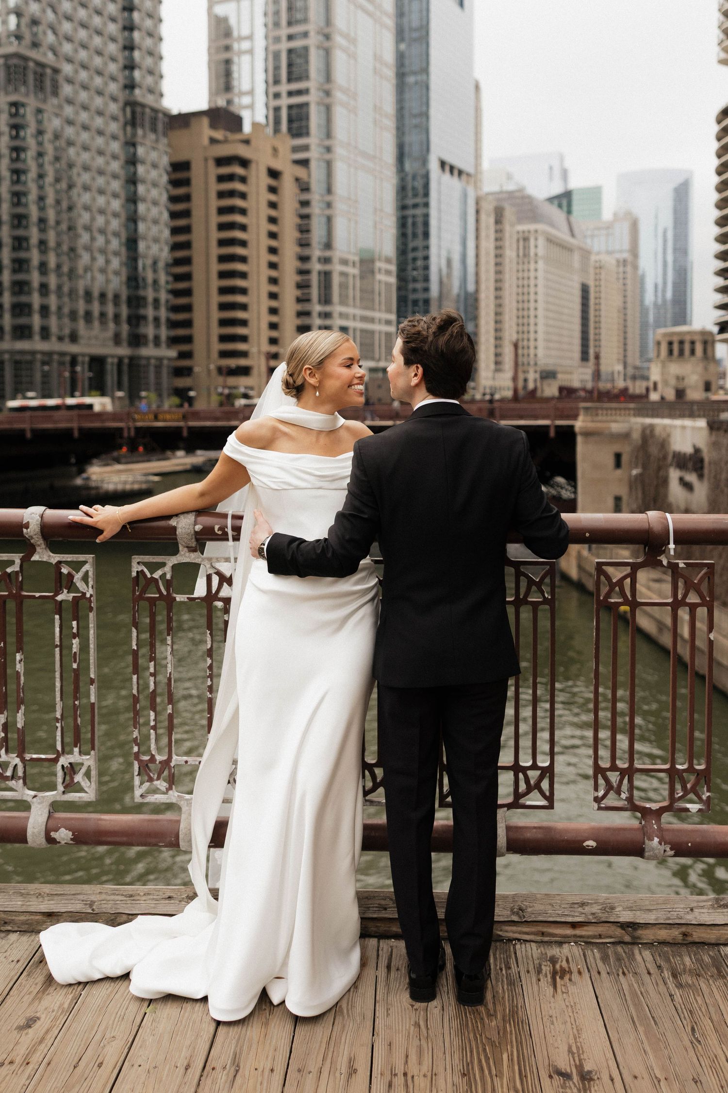 Bride and groom embracing on a bridge overlooking the Chicago River with city skyline backdrop.