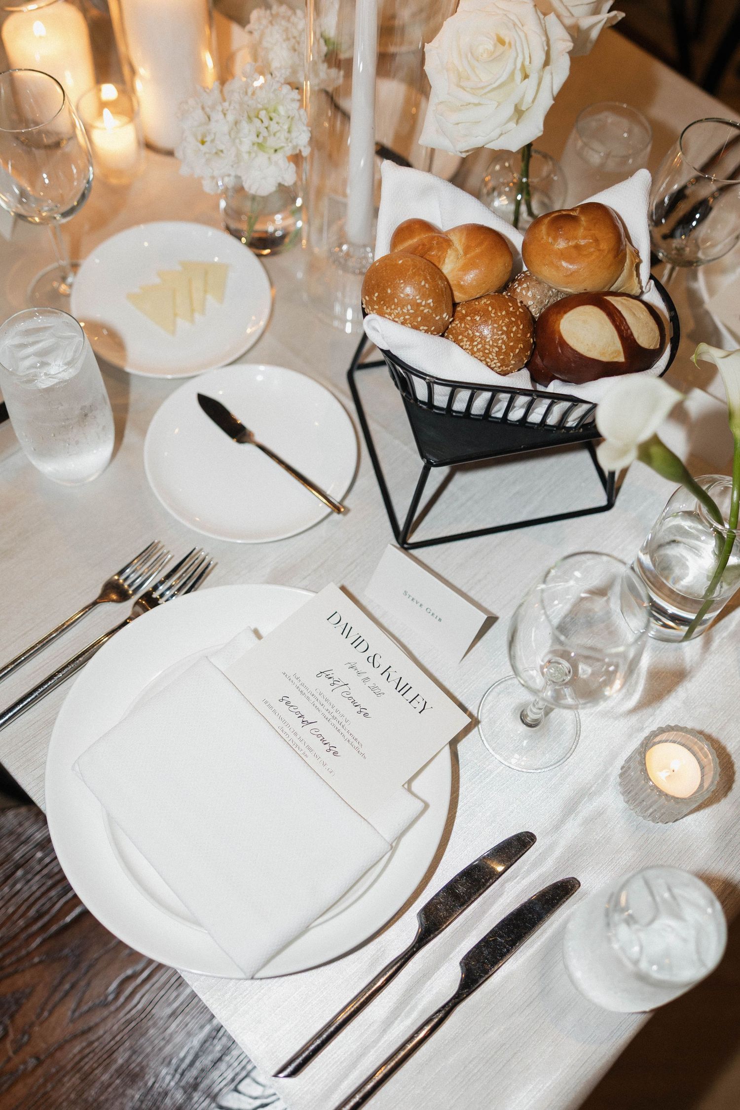 Elegant wedding table setting with white plates, gold cutlery, dinner rolls, candles, and personalized menu cards.