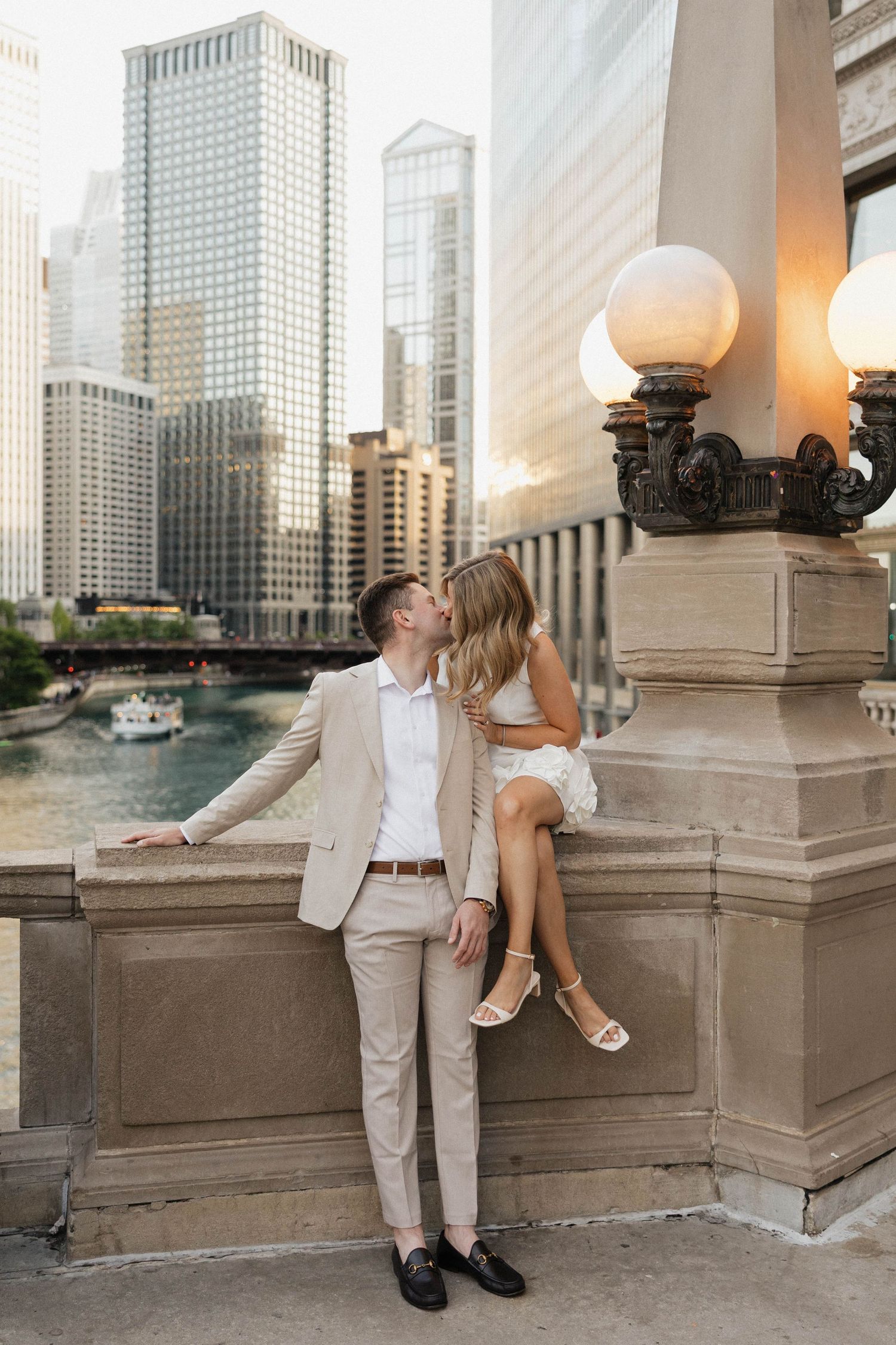 Couple sharing a romantic kiss on a Chicago Riverwalk bridge, man in beige suit, woman in white dress, city skyline backdrop.