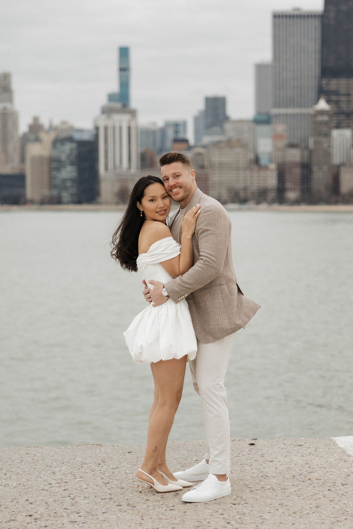 Couple embracing and smiling in white dress and tan blazer with Chicago skyline.