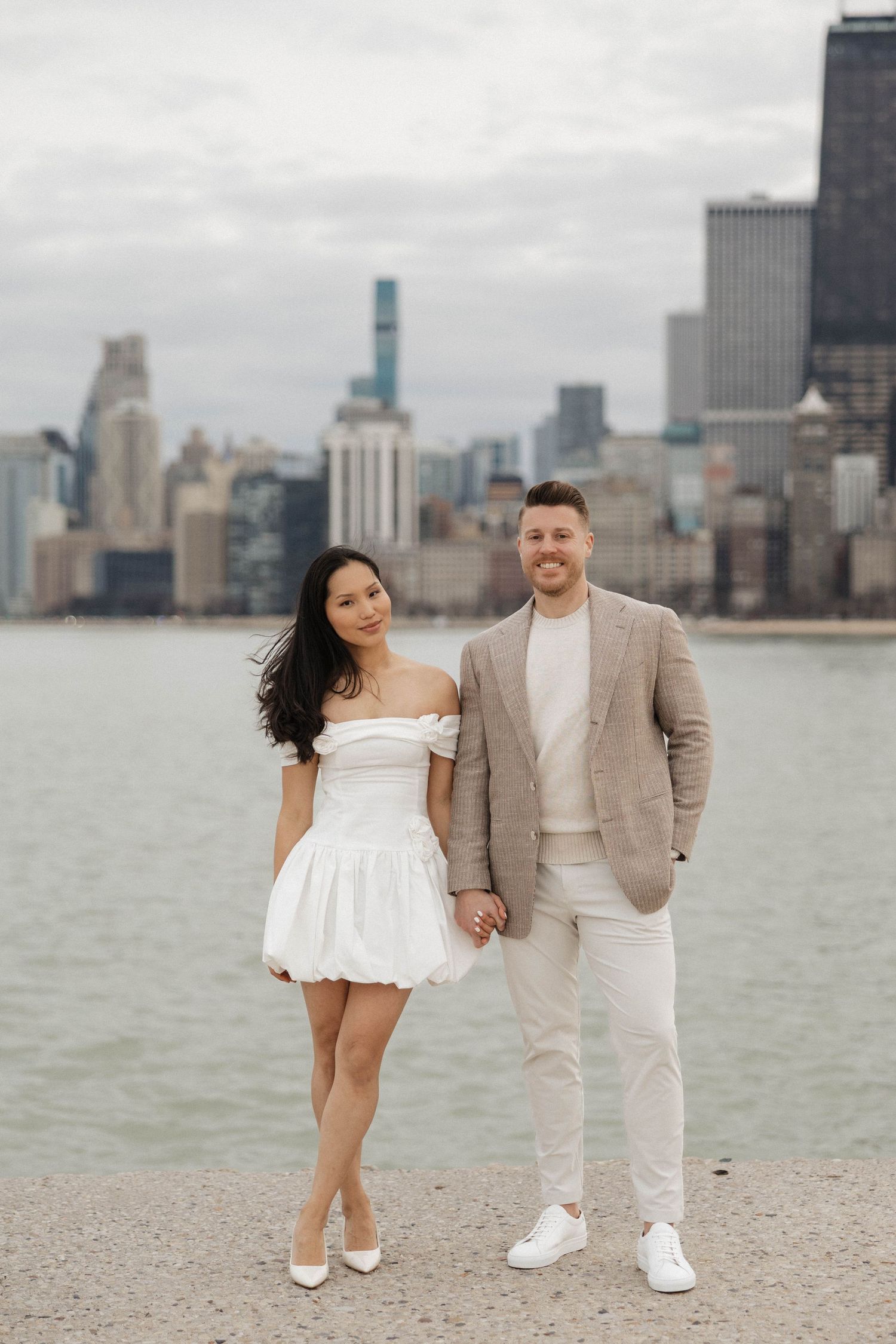Couple holding hands and smiling by Chicago lakefront skyline, woman in white mini dress, man in tan blazer.